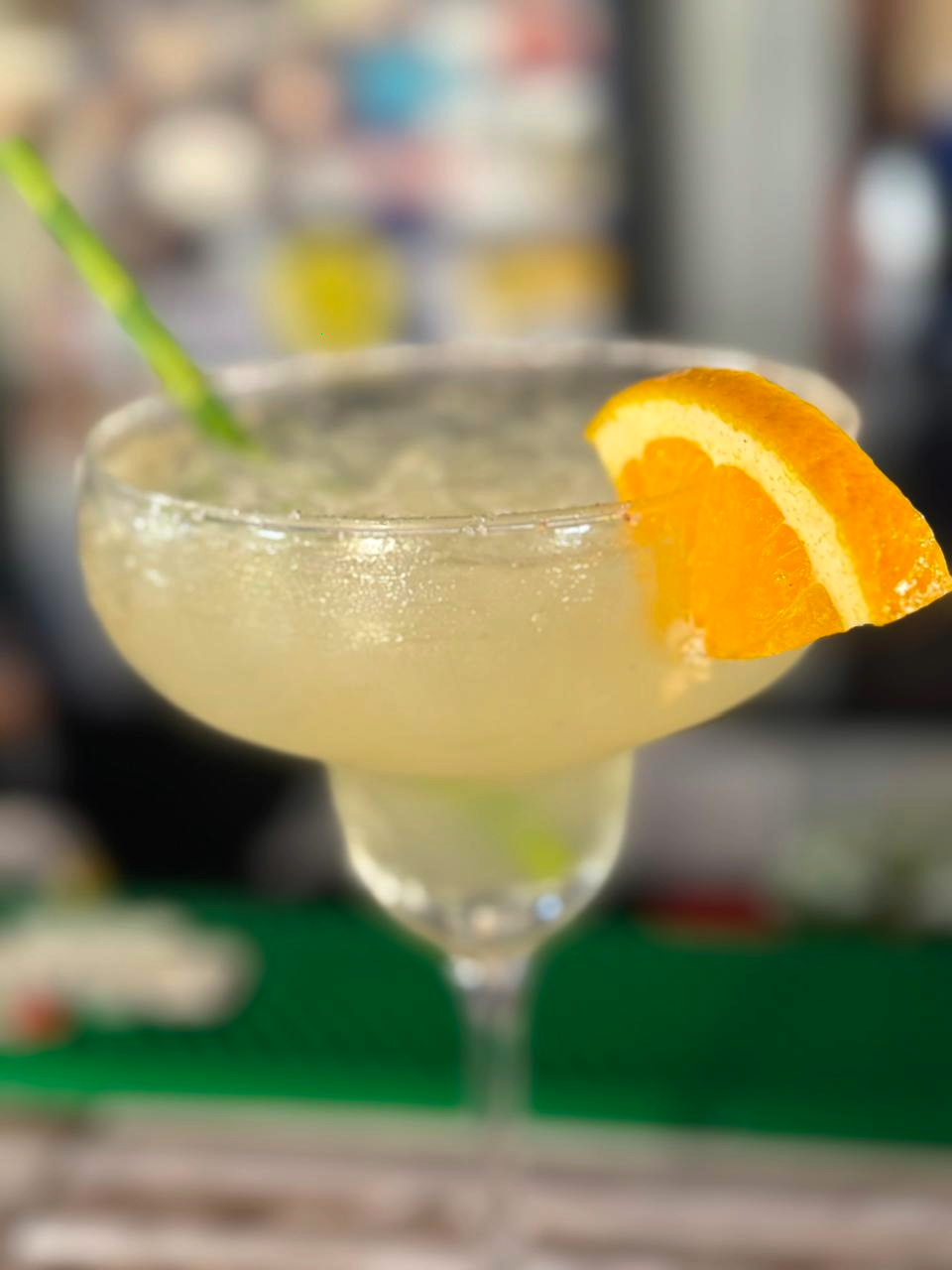 A cocktail with a salted rim, garnished with an orange slice and a green straw, on a bar counter.