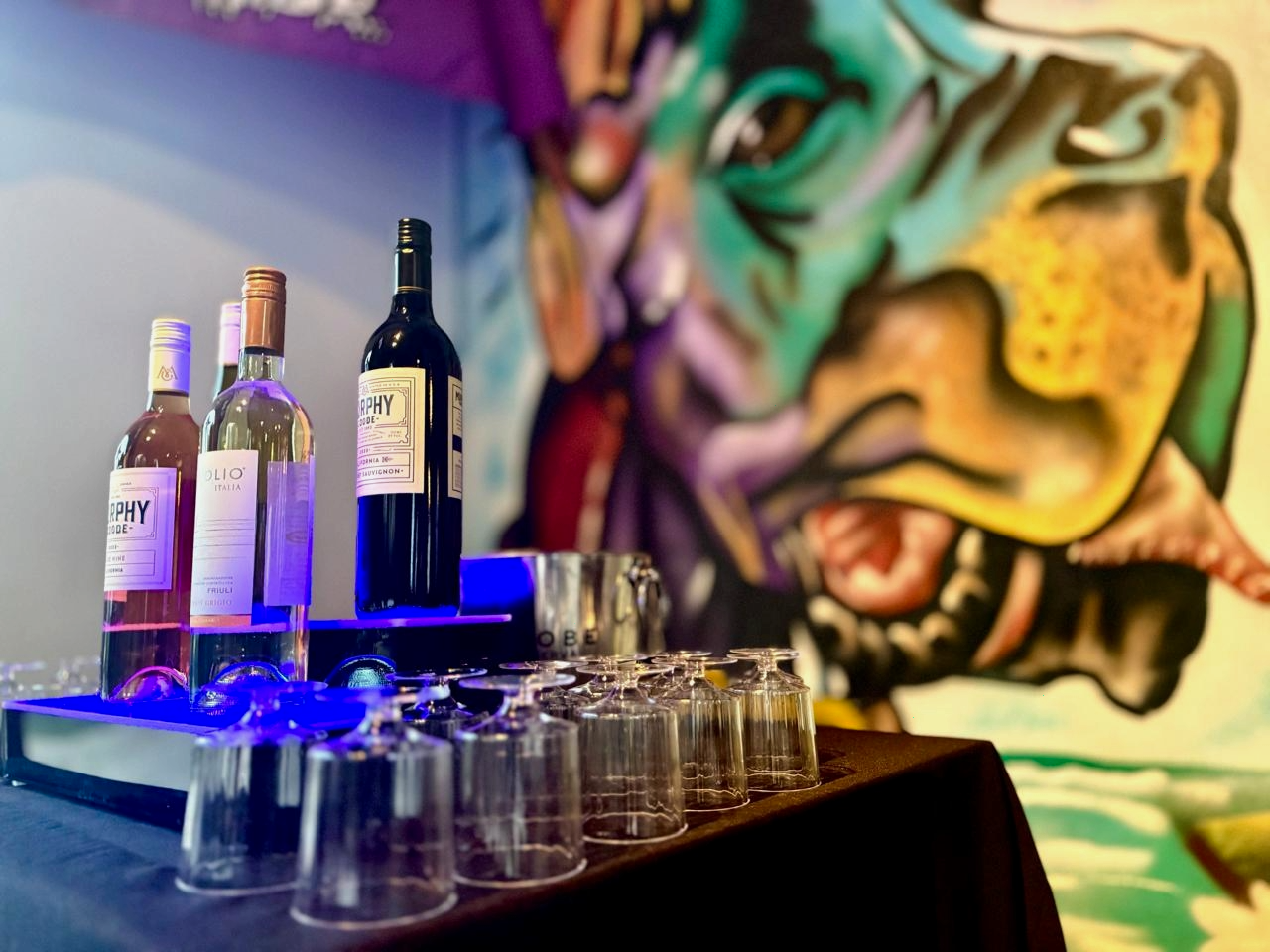 Bottles of wine and empty upside-down glasses on a table with a colorful abstract mural in the background.