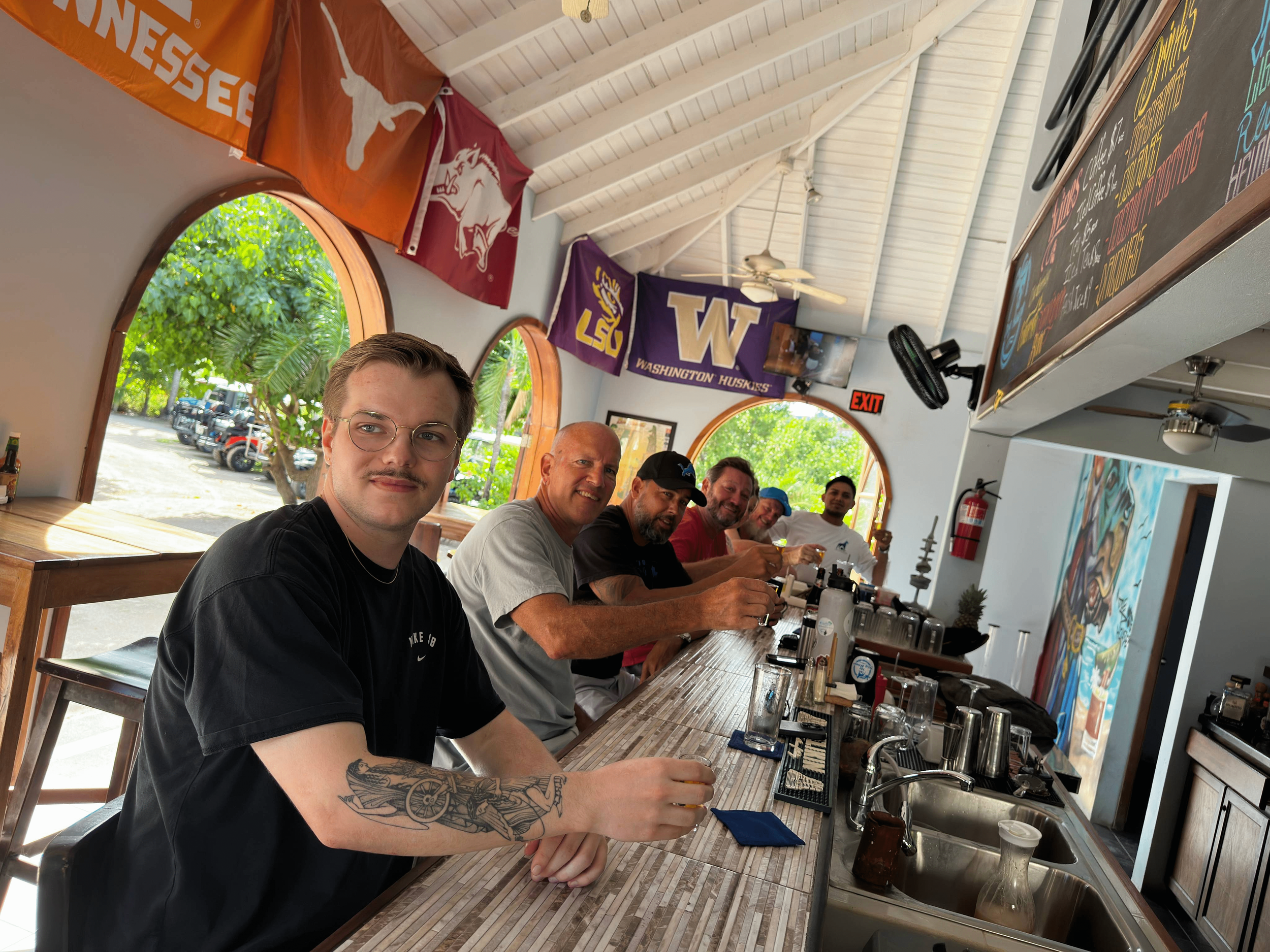 Six men sitting at a bar counter in a restaurant or cafe, smiling and enjoying drinks. The background features large windows with greenery outside, colorful flags hanging from the ceiling, and a chalkboard menu.
