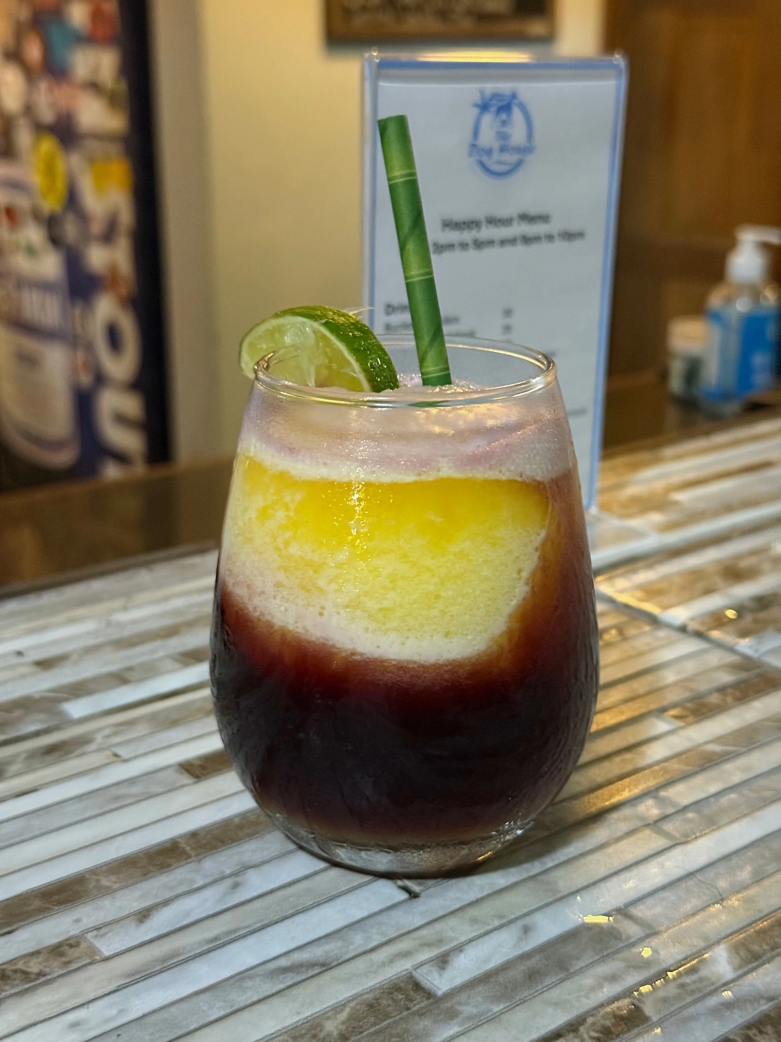 A cocktail with dark red or purple liquid, garnished with a lime wedge and a green straw, on a striped counter in a bar or restaurant.
