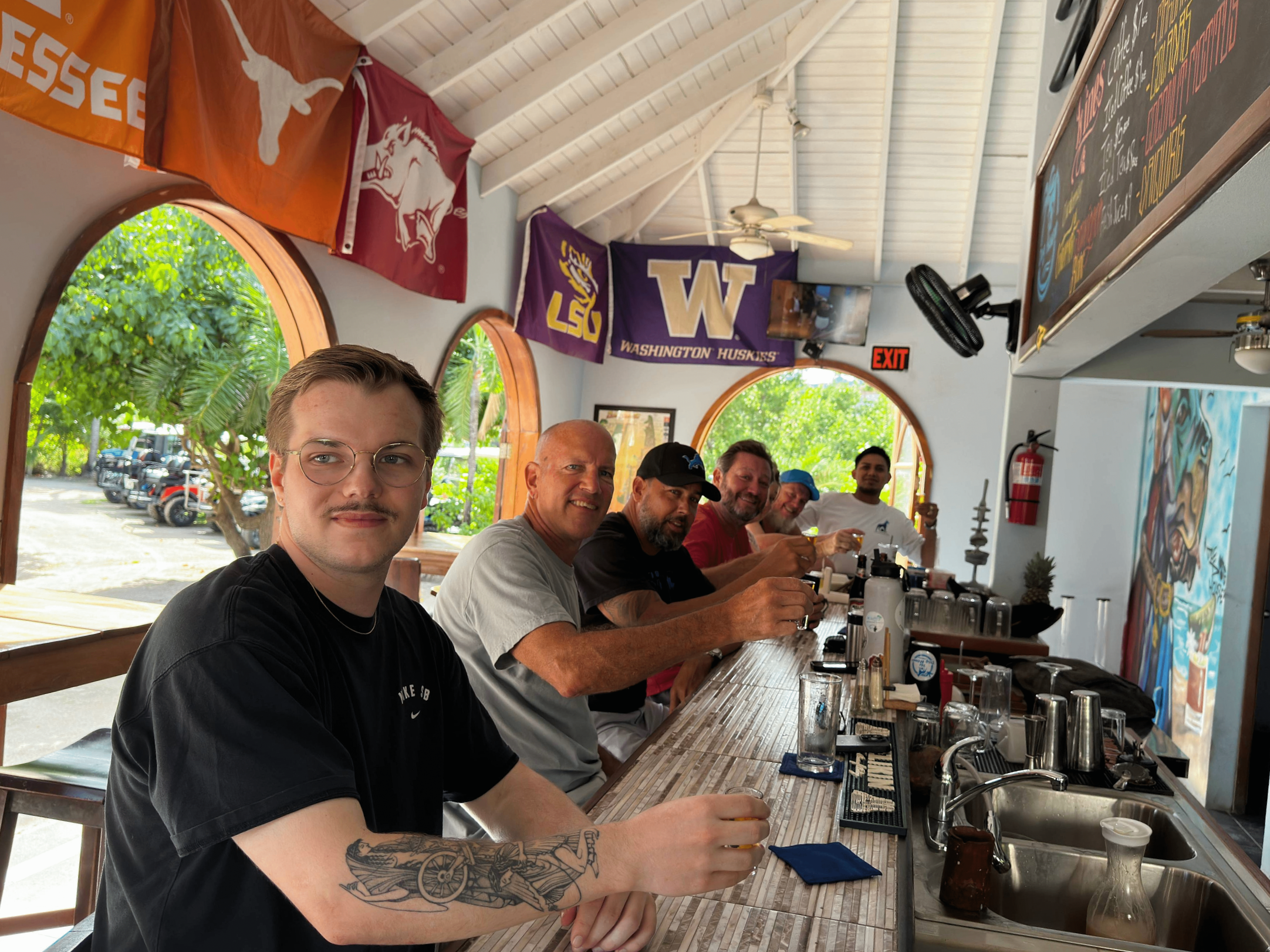 Group of men sitting at a bar counter in a restaurant, raising glasses, with flags of universities and sports teams hanging on the wall behind them.