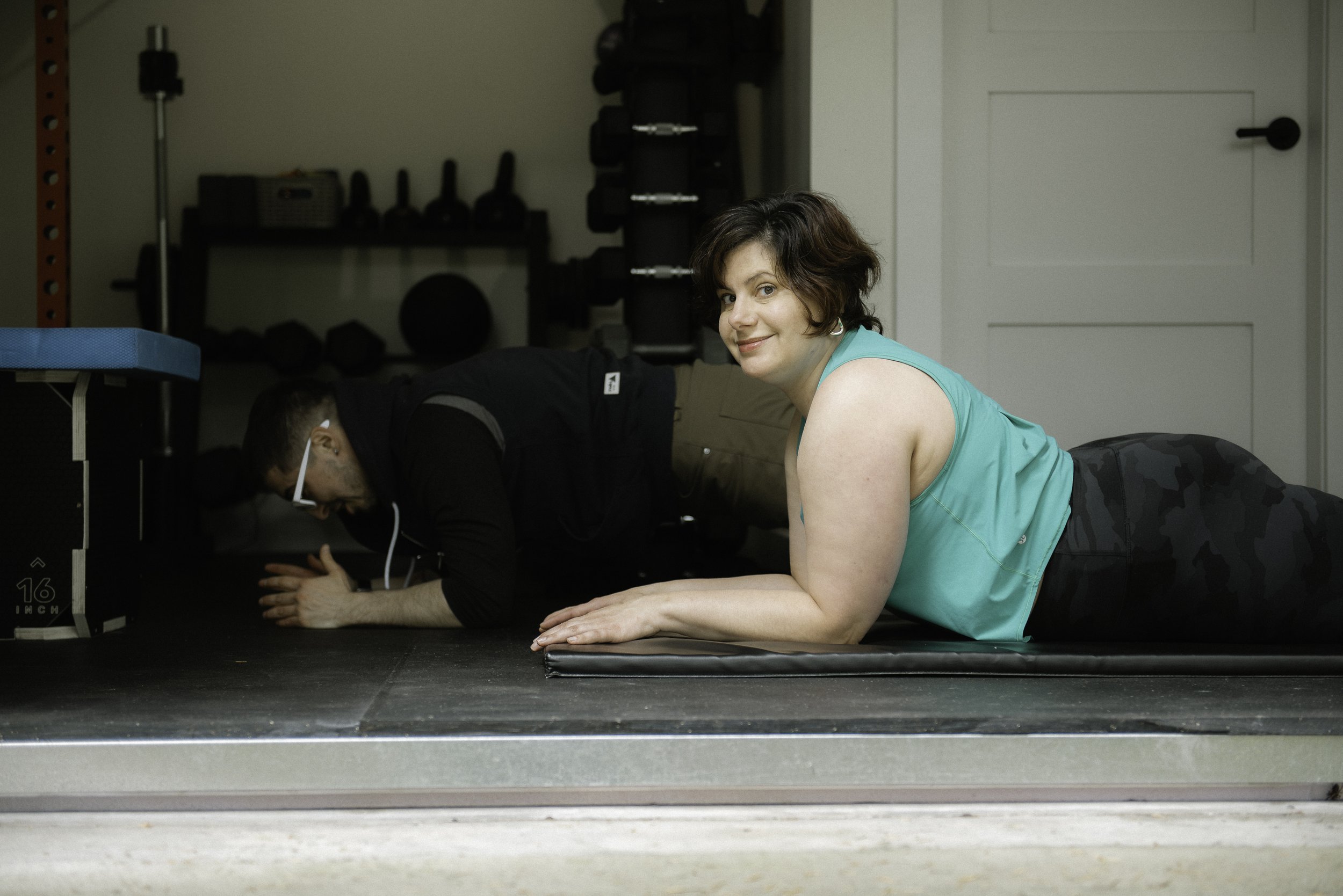 A woman in a green sports top is on a fitness mat smiling towards the camera