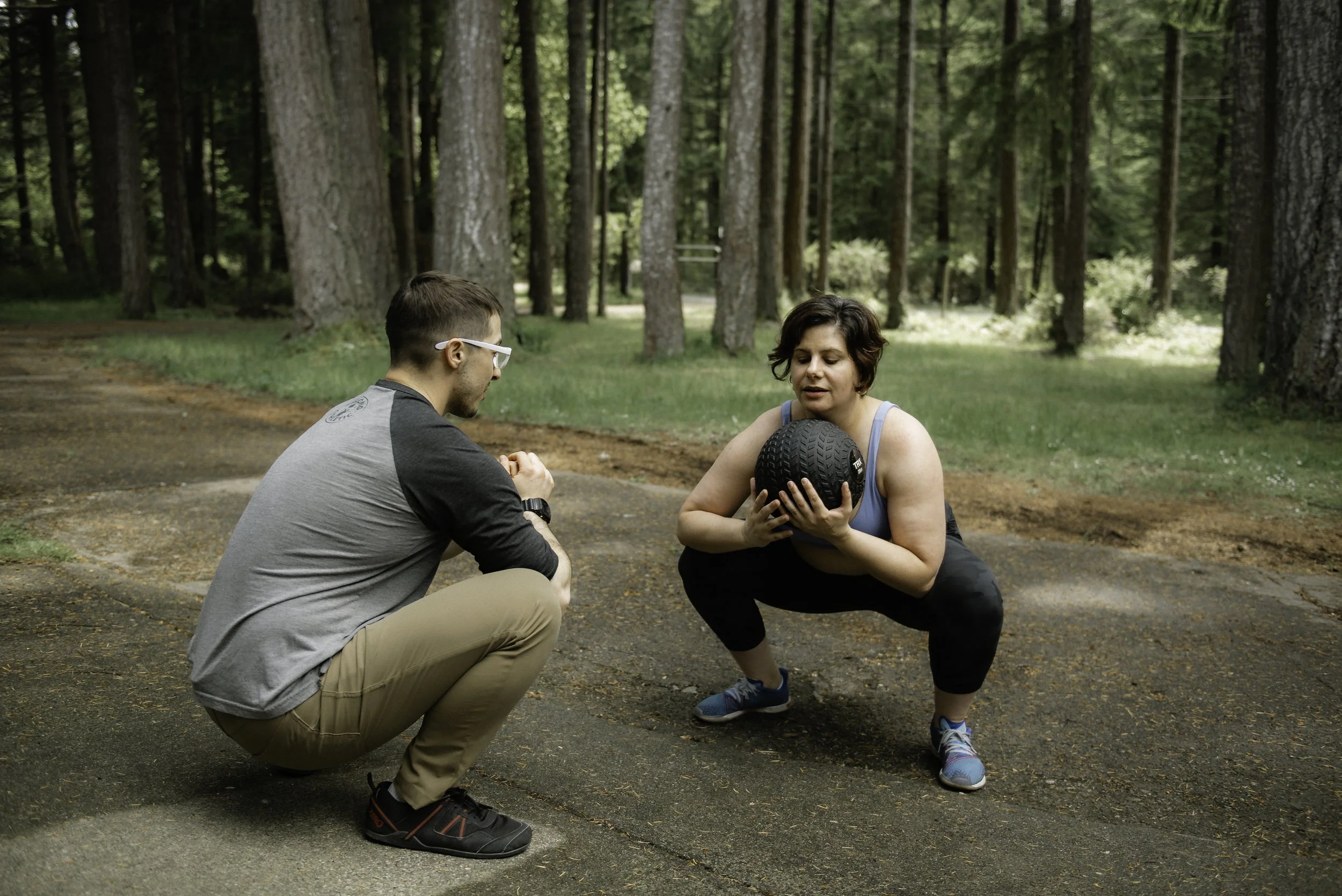 A man with tattoos, red glasses, and a sleeveless shirt stretching his arms upwards on outdoor fitness equipment in a park with green trees and benches.