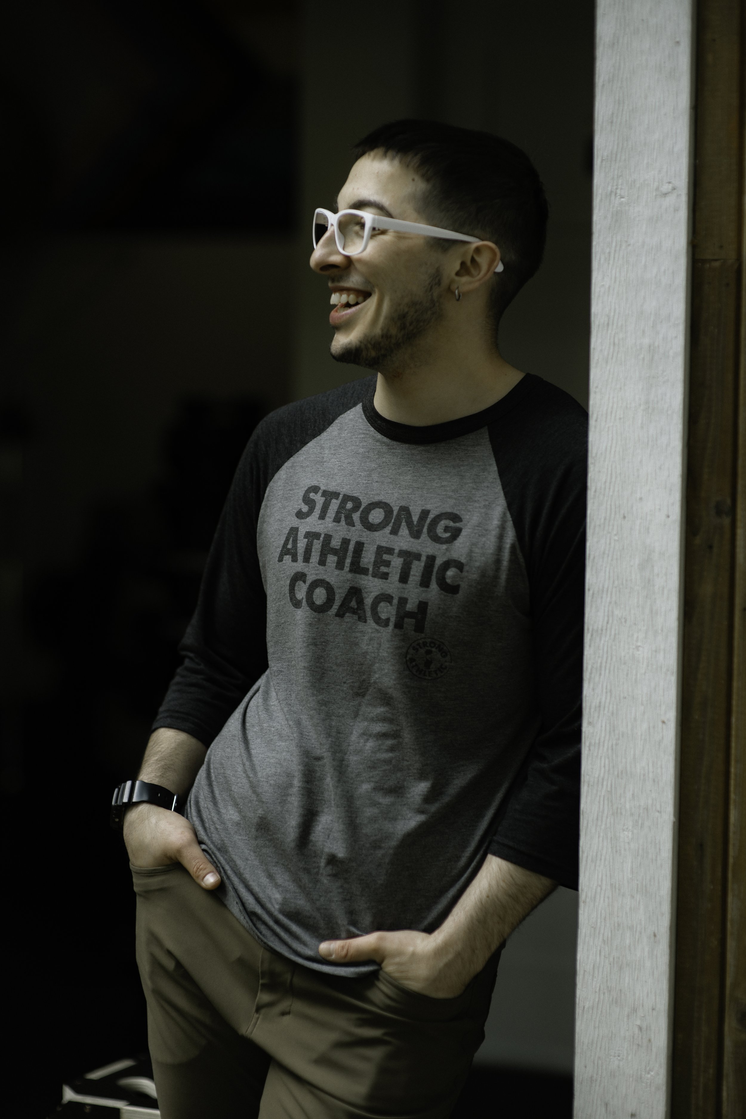 Dibs stands in side profile, smiling warmly, wearing a shirt that says Strong Athletic Coach.