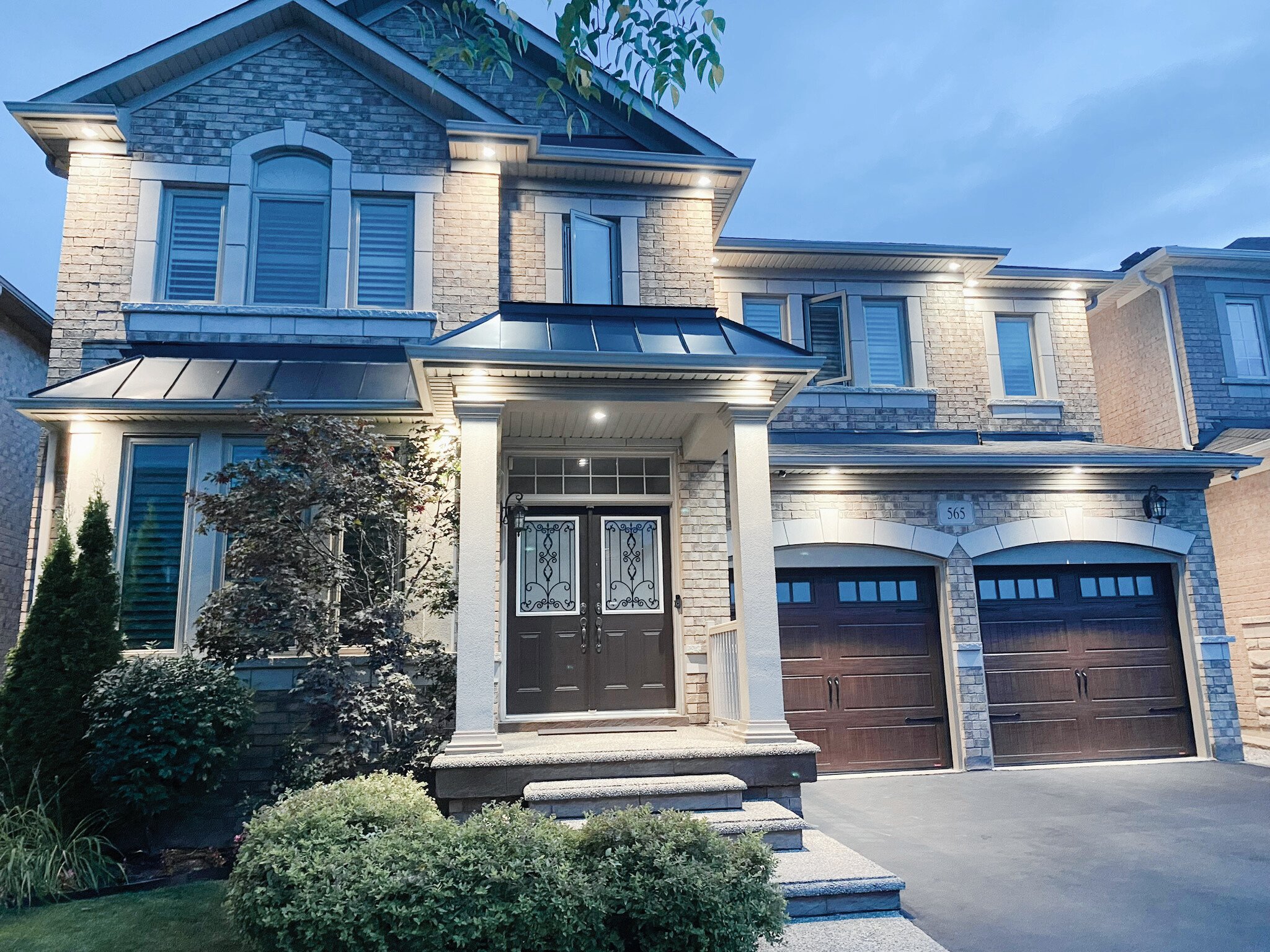 Two-story brick house with decorative double doors and two-car garage, surrounded by manicured landscaping, and illuminated by exterior lights in the evening.