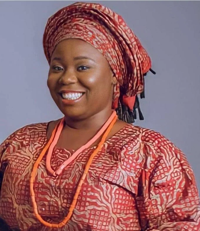 A woman wearing traditional African attire with a headwrap and beaded necklace, smiling at the camera.