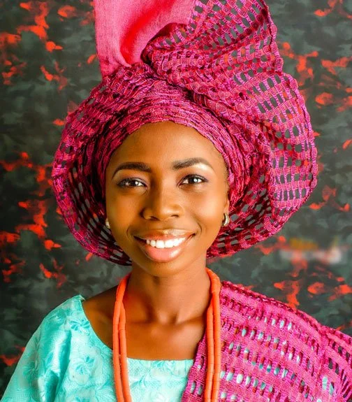 Young woman wearing traditional colorful attire and headwrap, smiling against a dark background.