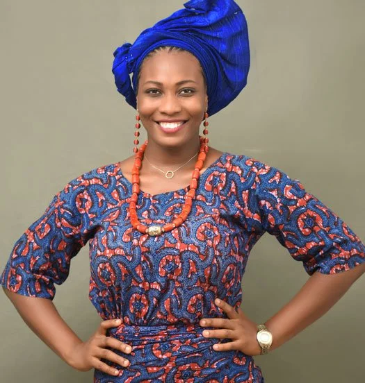 A smiling woman wearing a blue and red patterned dress, a matching blue headwrap, large red bead necklace, smaller pendant necklace, and a watch, standing against a gray background.