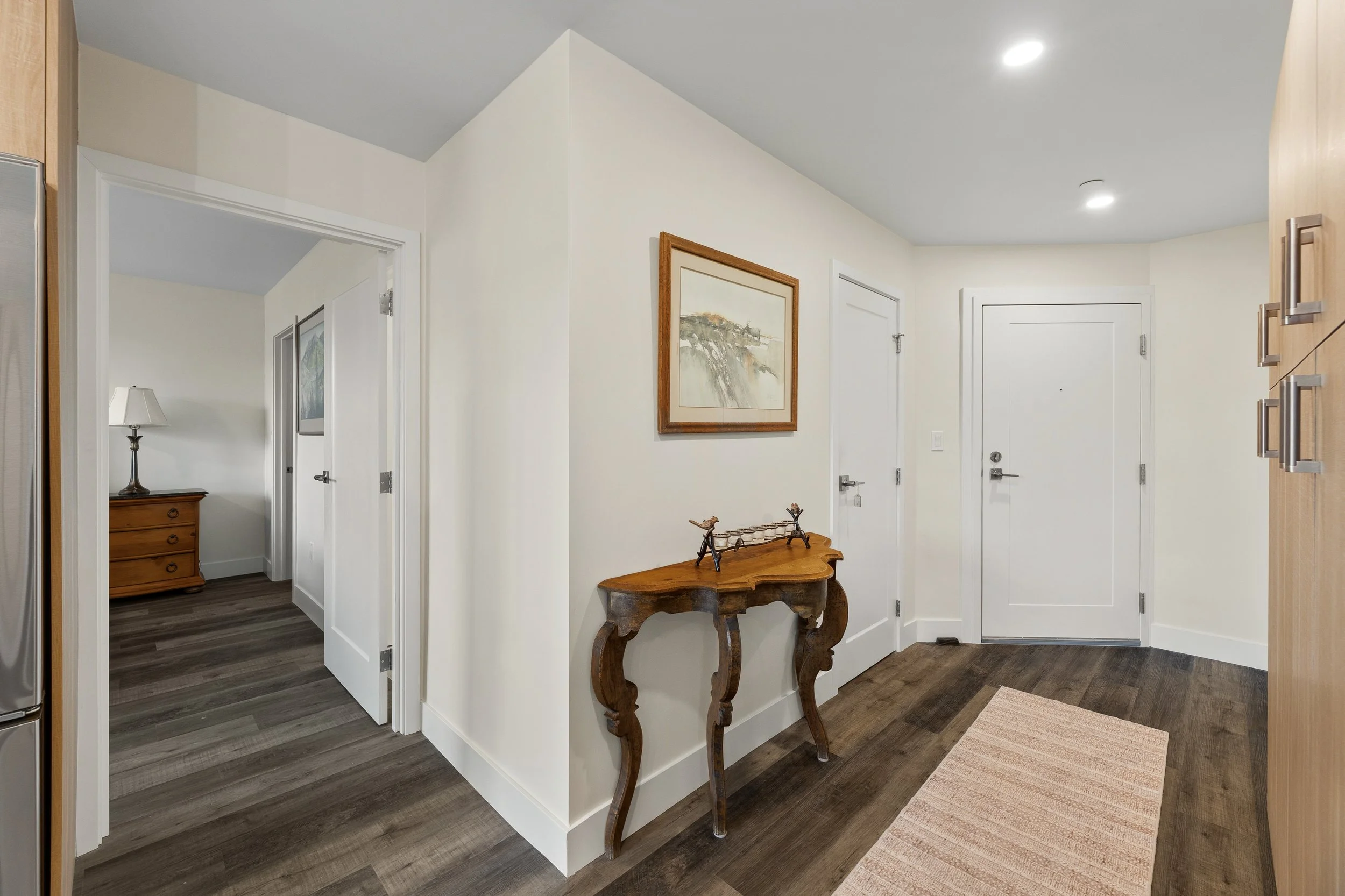 Interior view of a modern home entryway with a small wooden table, artwork on the wall, and doors leading to other rooms.