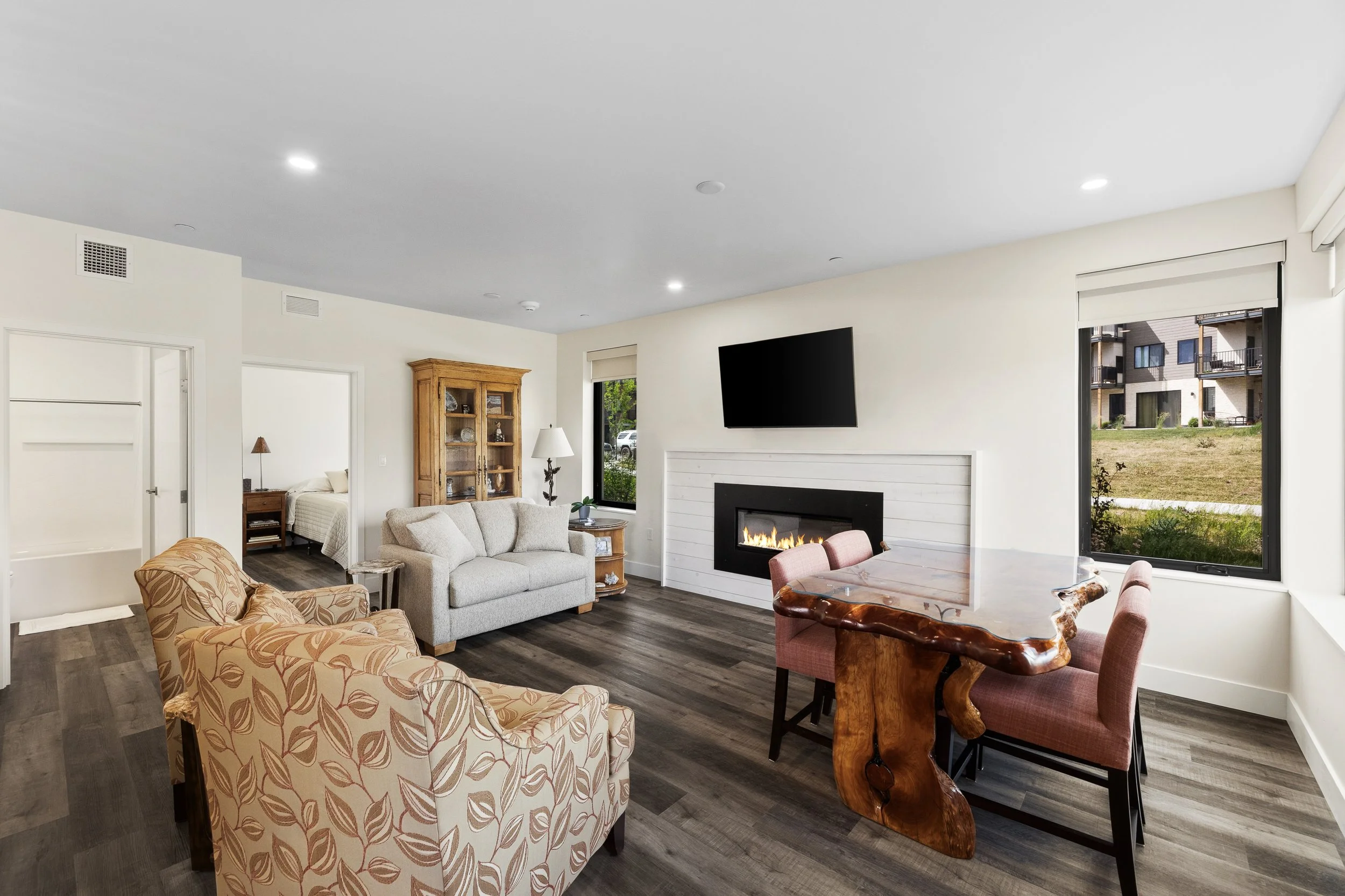 Living room with beige and patterned armchairs, a gray sofa, a wooden dining table with chairs, a TV above a fireplace, large windows showing an outdoor building and grass, wooden flooring, and white walls.