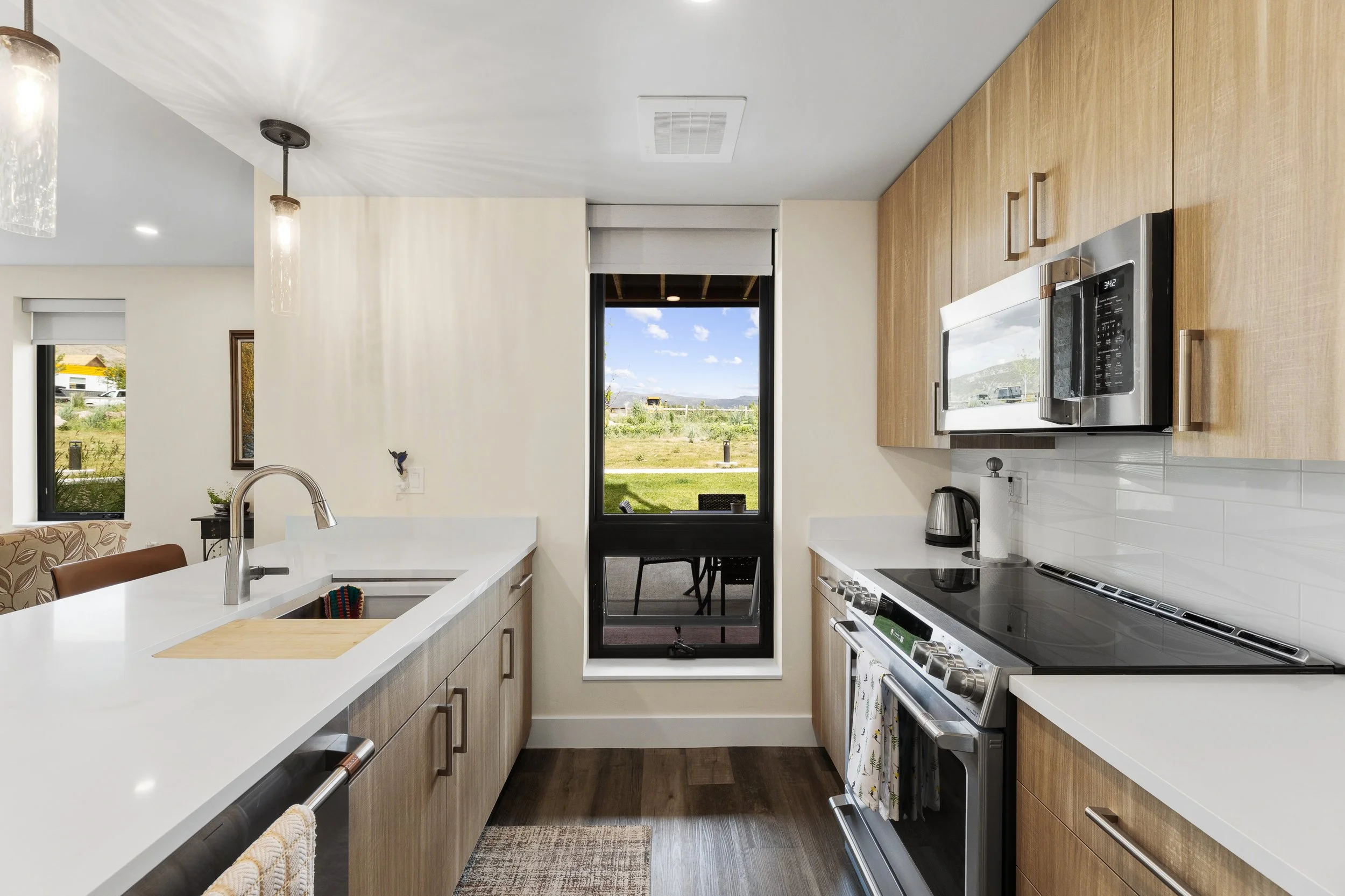 Modern kitchen with white countertops, wooden cabinets, stainless steel appliances, a window and a door leading outside, with a view of greenery and blue sky.