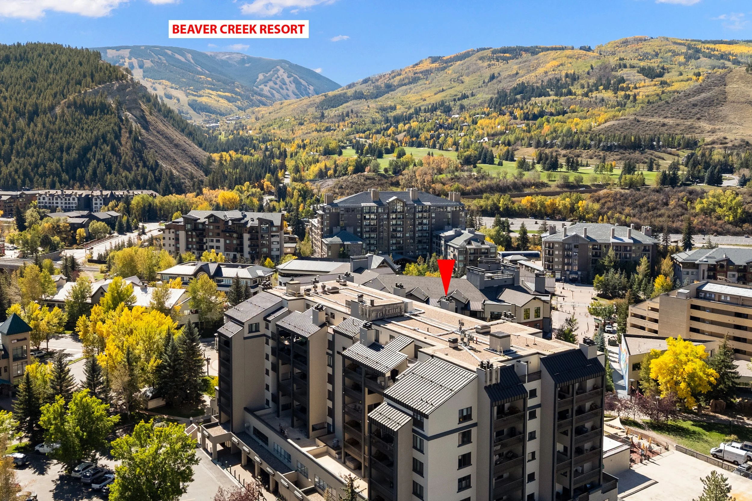 A view of the mountains with ski slopes, a residential area with trees and apartment buildings, and a part of the Beaver Creek Resort, highlighted by a red arrow.