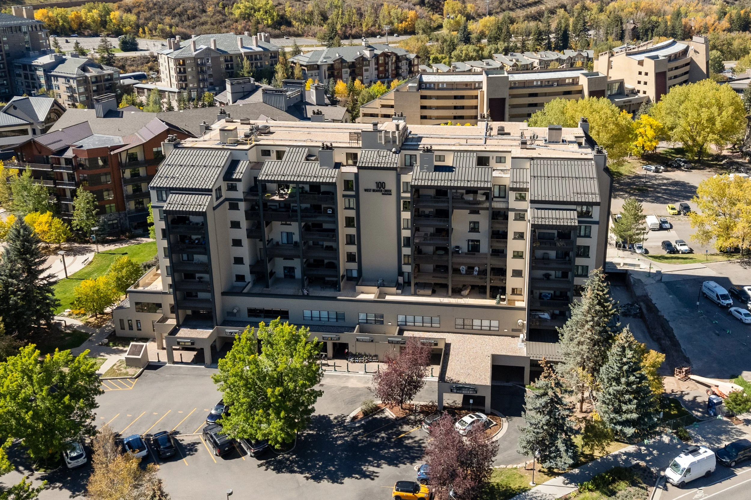 Aerial view of a modern apartment complex surrounded by green trees with some fall foliage, parking lots, and additional residential buildings in the background, set amid a hilly landscape.