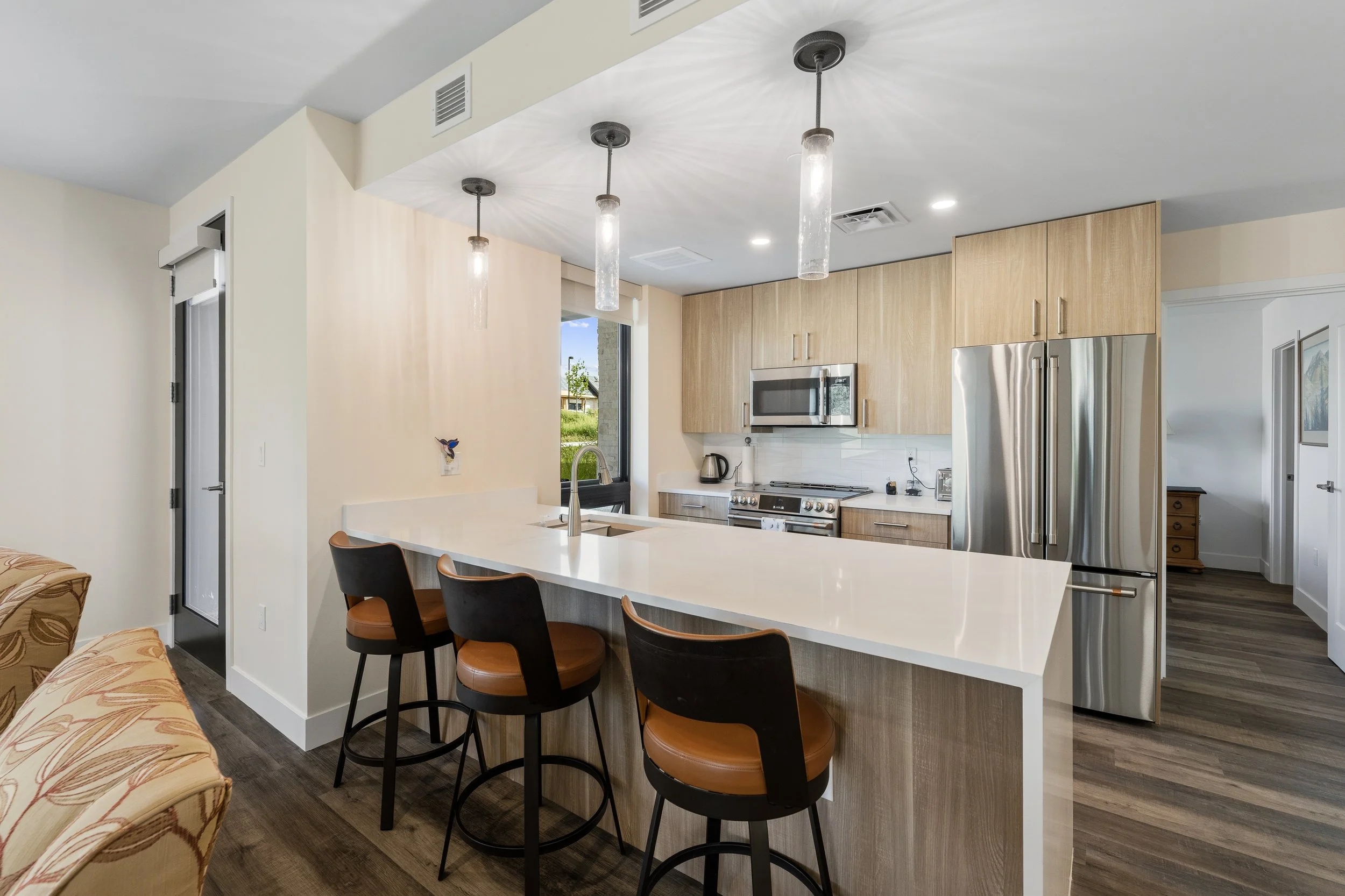 Modern kitchen with wooden cabinets, stainless steel appliances, white countertops, pendant lights, and a breakfast bar with black and brown chairs.