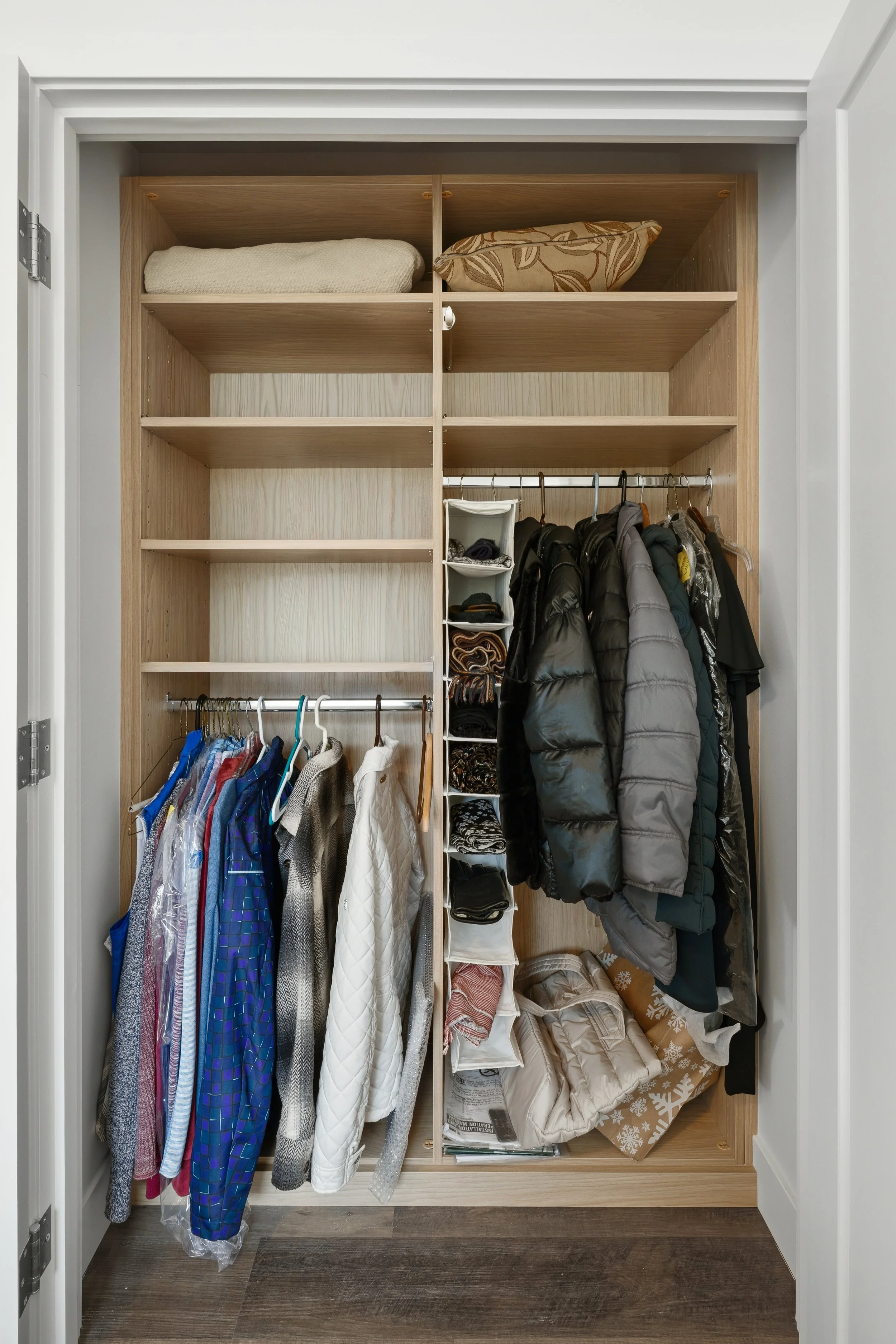 A closet with wooden shelves and a hanging rod with jackets and shirts, some wrapped in plastic. The top shelf has pillows and blankets, while the bottom includes storage boxes, folded clothes, and a wrapped gift bag.