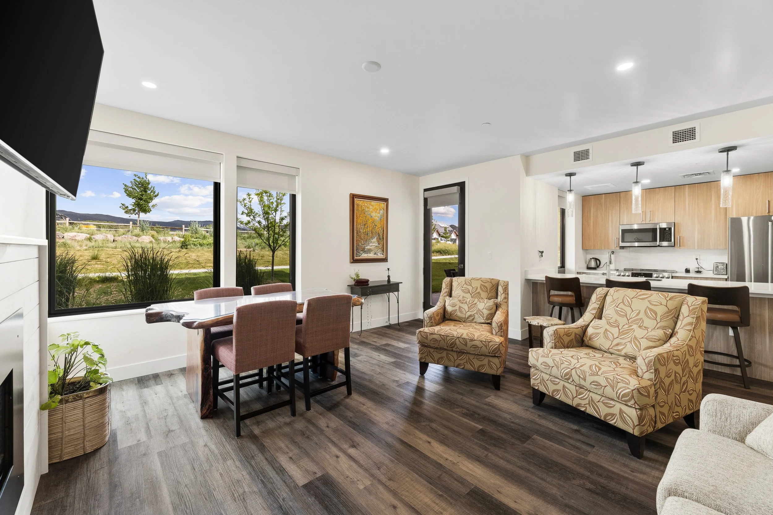Interior view of a modern living room and kitchen area with large windows showing a green outdoor landscape, dining table with chairs, beige armchairs with floral pattern, and kitchen with wooden cabinets and stainless steel appliances.