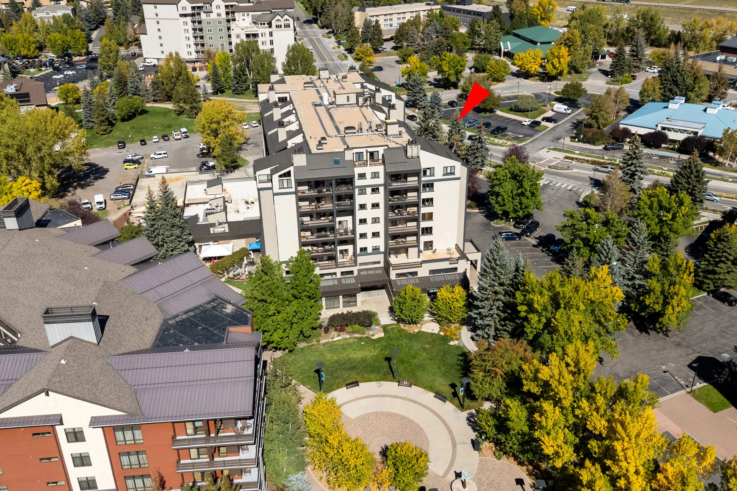 An aerial view of a residential area with multiple apartment buildings, parking lots, and a park with trees and a circular pathway.