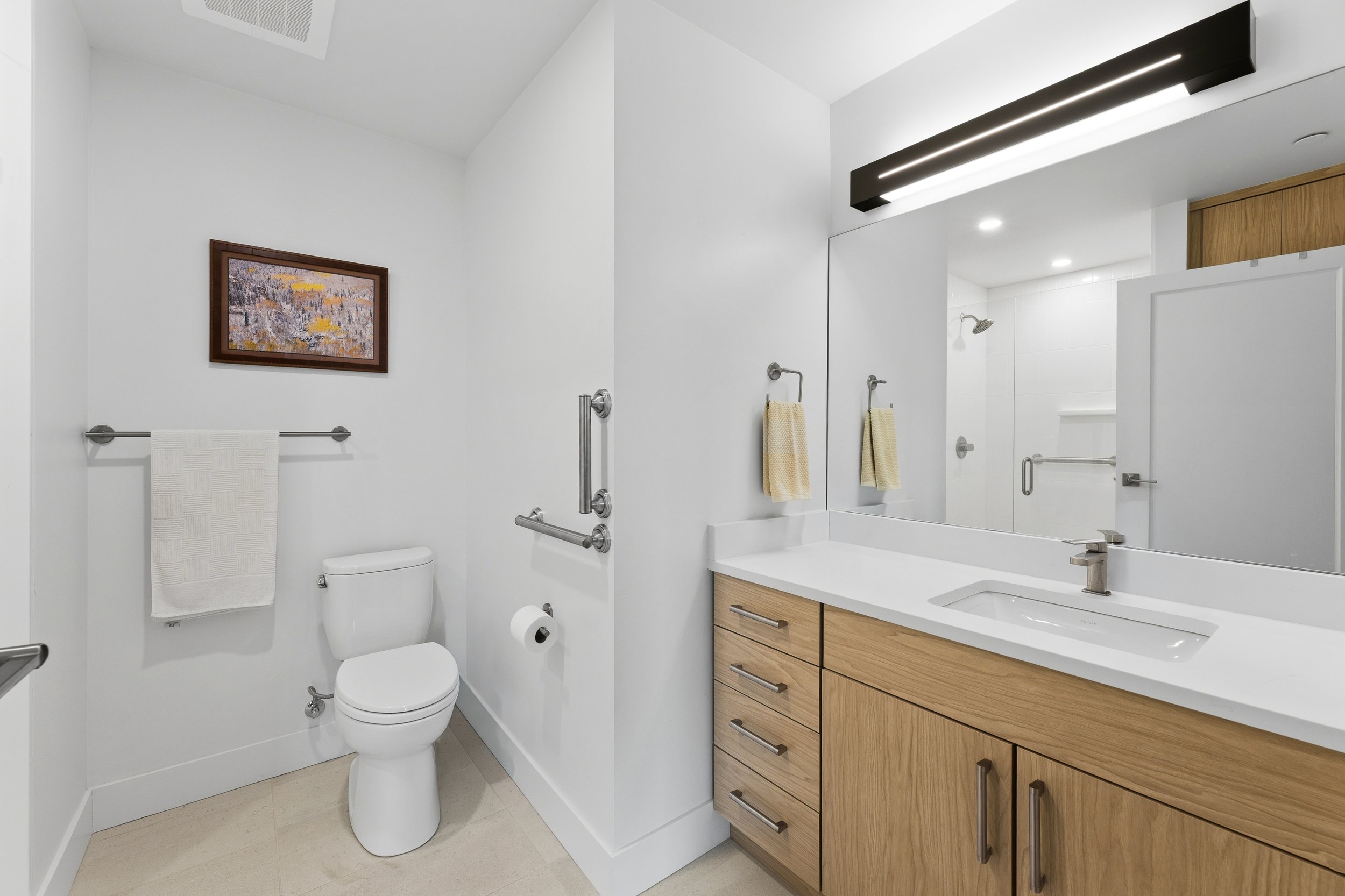 Modern bathroom featuring a toilet, a large mirror with a black lighting fixture above, a wooden vanity with drawers and a sink, yellow towels, a framed painting on the white wall, and a shower in the background.