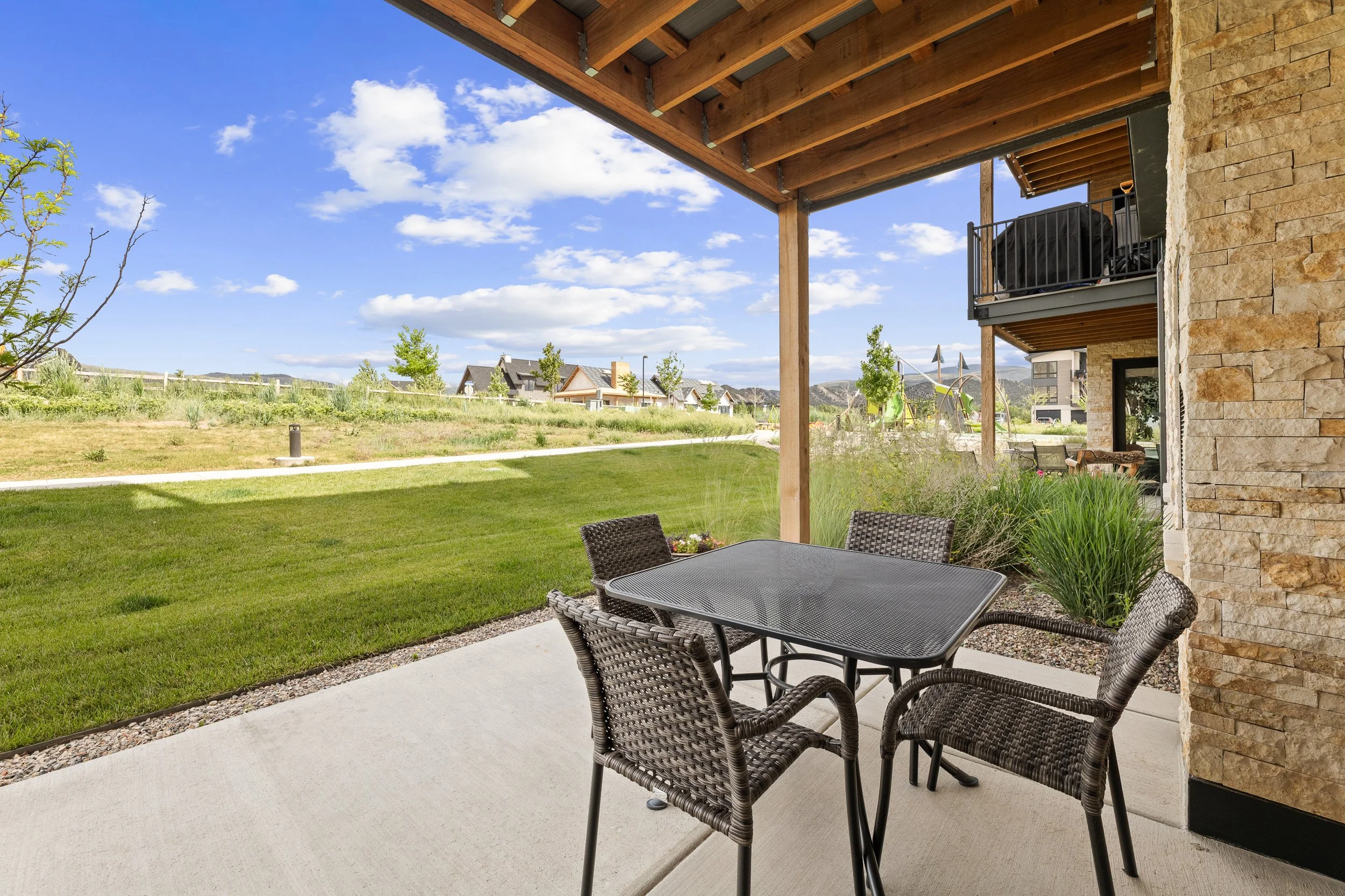 View of a backyard patio with a glass table and wicker chairs, overlooking a grassy yard with trees and houses under a blue sky with scattered clouds.