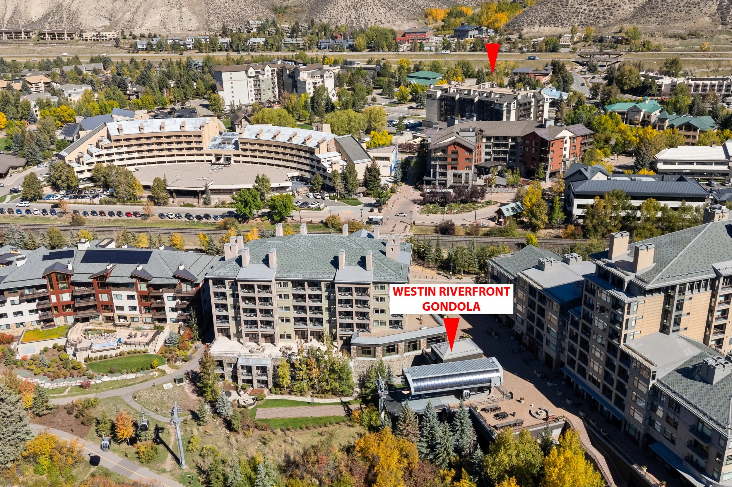 An aerial view of a city area with multiple buildings, trees, parking lots, and a cable car system with a gondola. There are mountains in the background. A red arrow points to a building with a green roof, and text overlay indicates 'Westin Riverfron