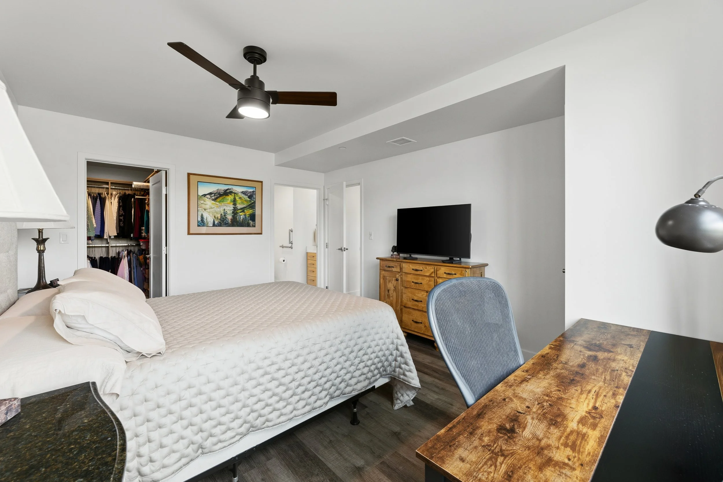 A bedroom with a bed, a desk with a chair, a wooden dresser with a television, a ceiling fan, and open closet doors with clothing inside.