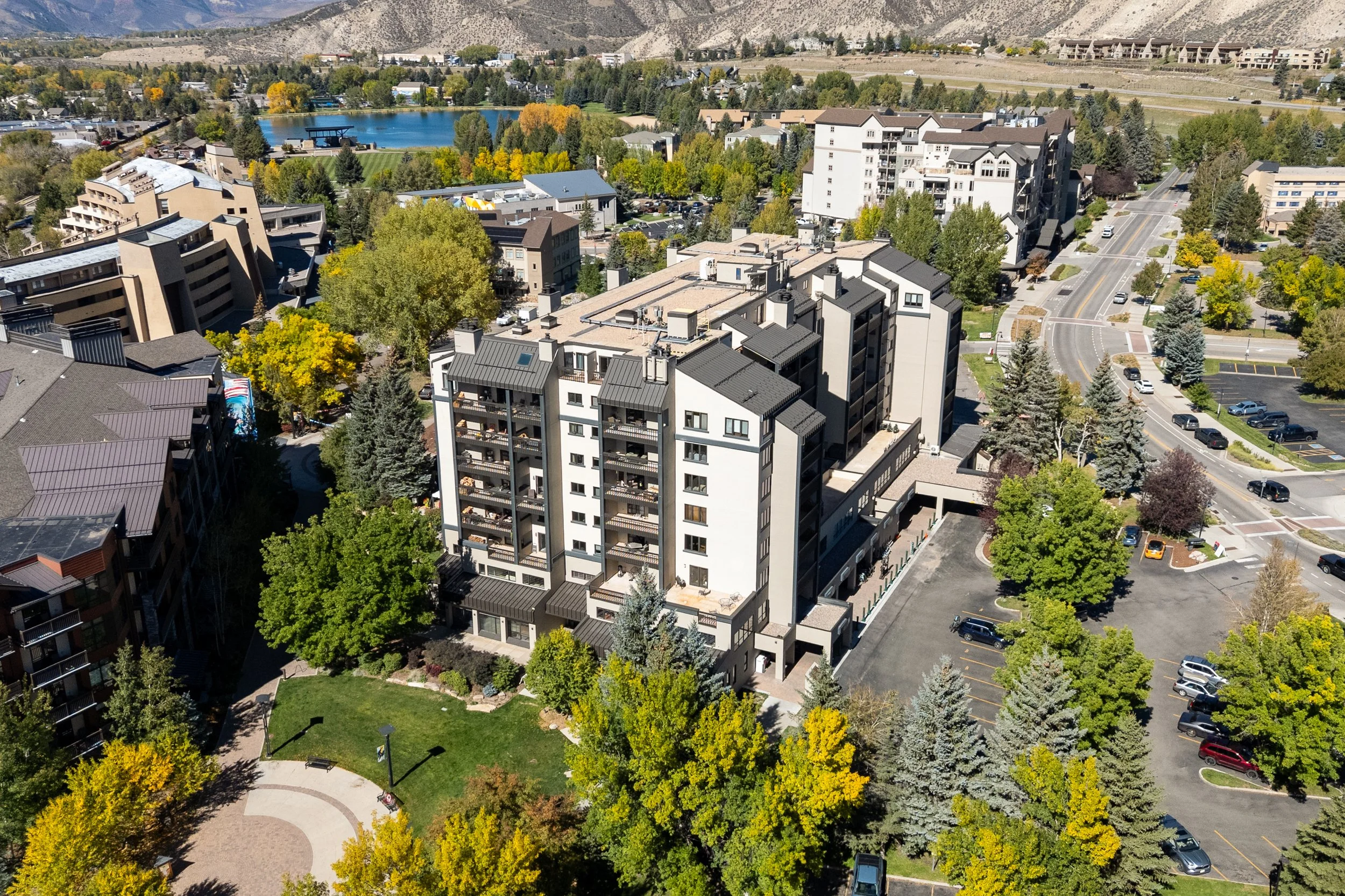 Aerial view of a city with residential buildings, a park with trees and a pond, parking lots, and mountain hills in the background.