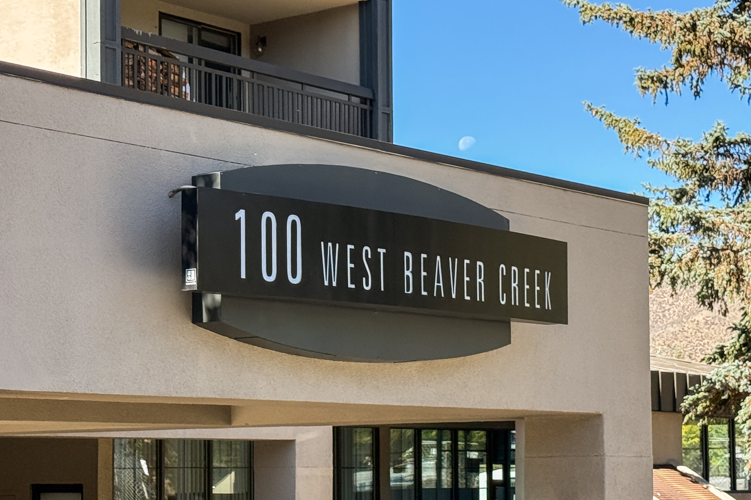 Building with a sign that reads '100 West Beaver Creek' and a balcony with a person on it, trees, and a clear blue sky with a visible moon.