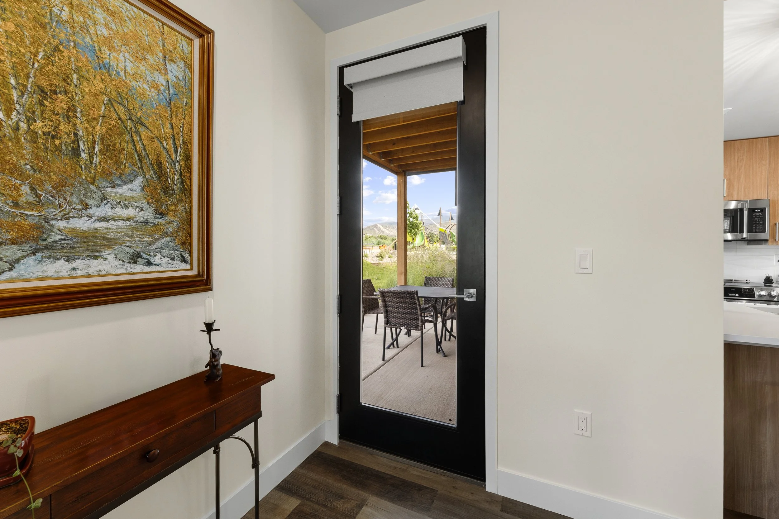 Interior view of a doorway leading to an outdoor patio area with a wooden pergola, a table, and chairs. Interior features include a framed painting of trees with orange leaves, a wooden console table with a small decorative object, and part of a kitc