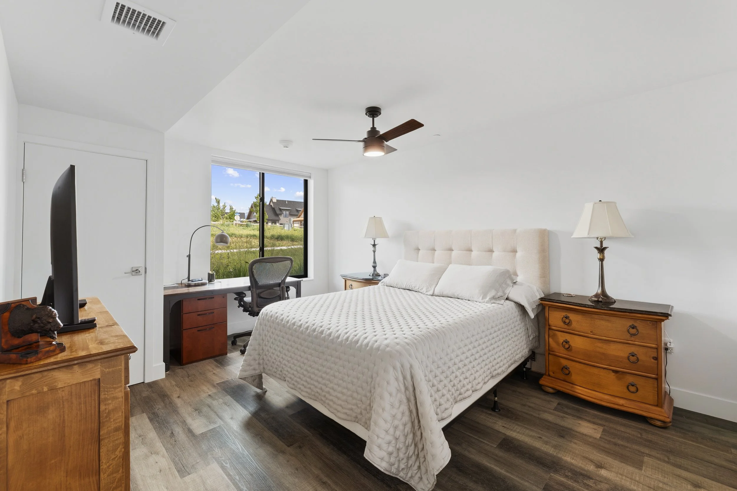 A bedroom with a white bed, beige headboard, and two wooden nightstands with lamps. There is a window showing a neighborhood outside and a ceiling fan.