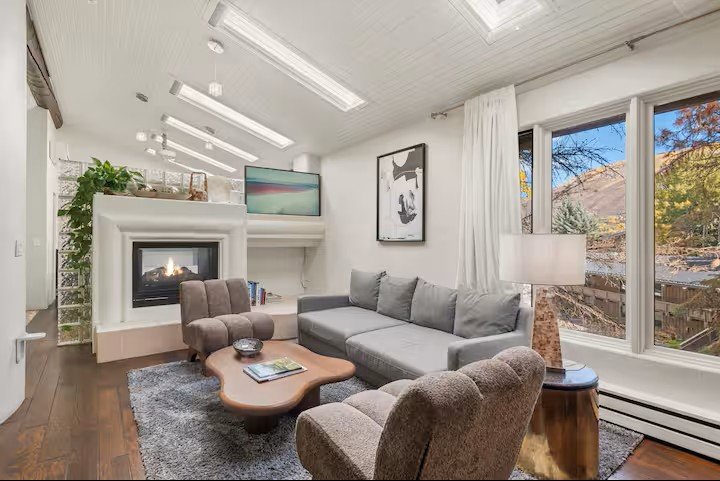 Living room with gray sofa, two brown armchairs, wooden coffee table, gray rug, stone fireplace, large window with white curtains, and a landscape view outside.