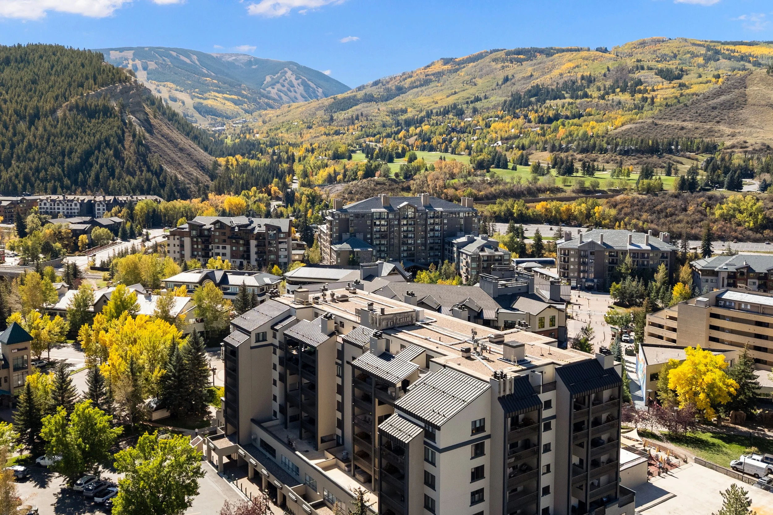 Aerial view of a mountain town with modern apartment buildings surrounded by trees with fall foliage, and forested mountains in the background under a blue sky.
