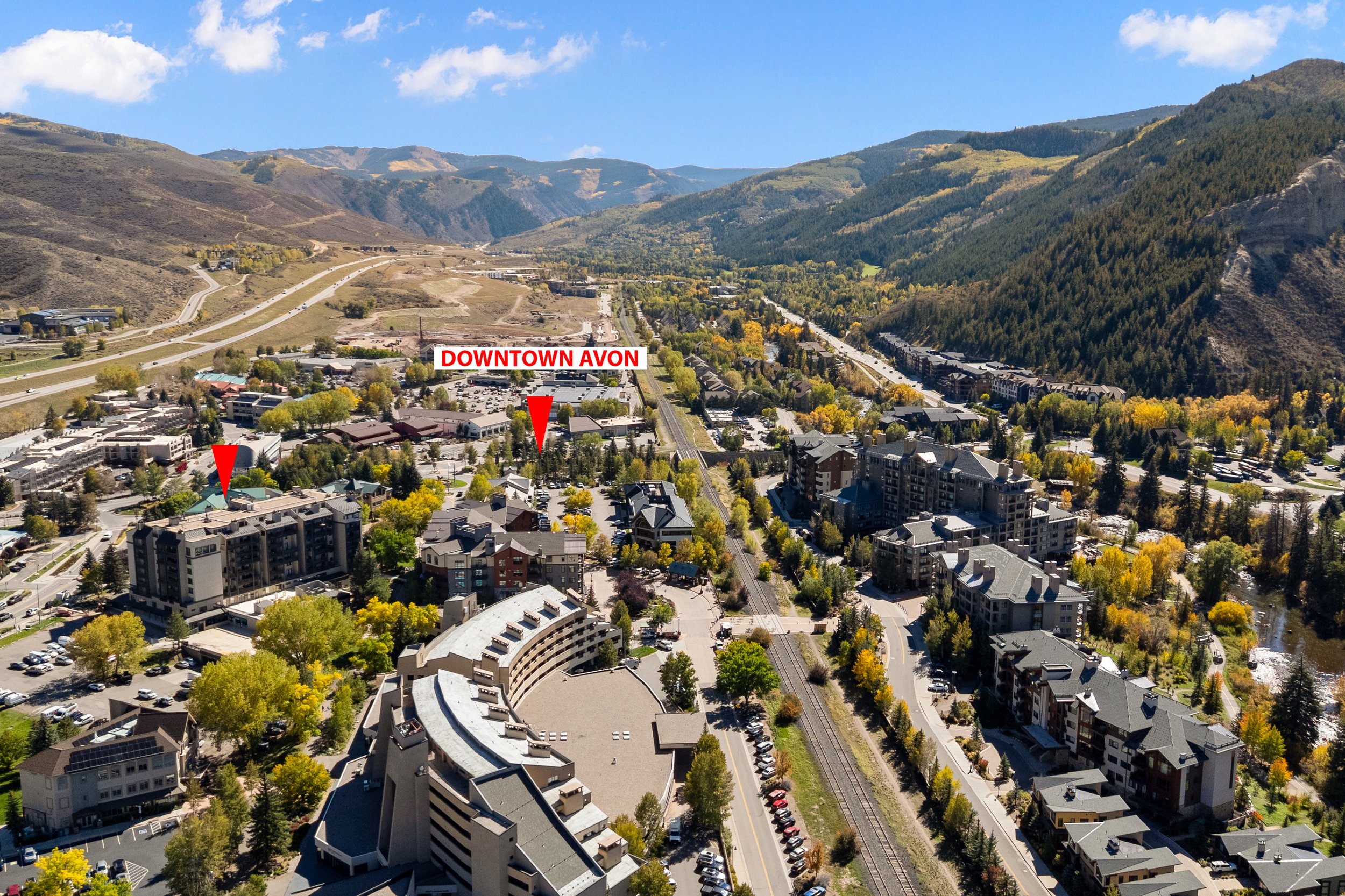 Aerial view of downtown Avon, Colorado, with a mix of residential and commercial buildings, surrounded by mountain ranges and greenery, under a partly cloudy sky.