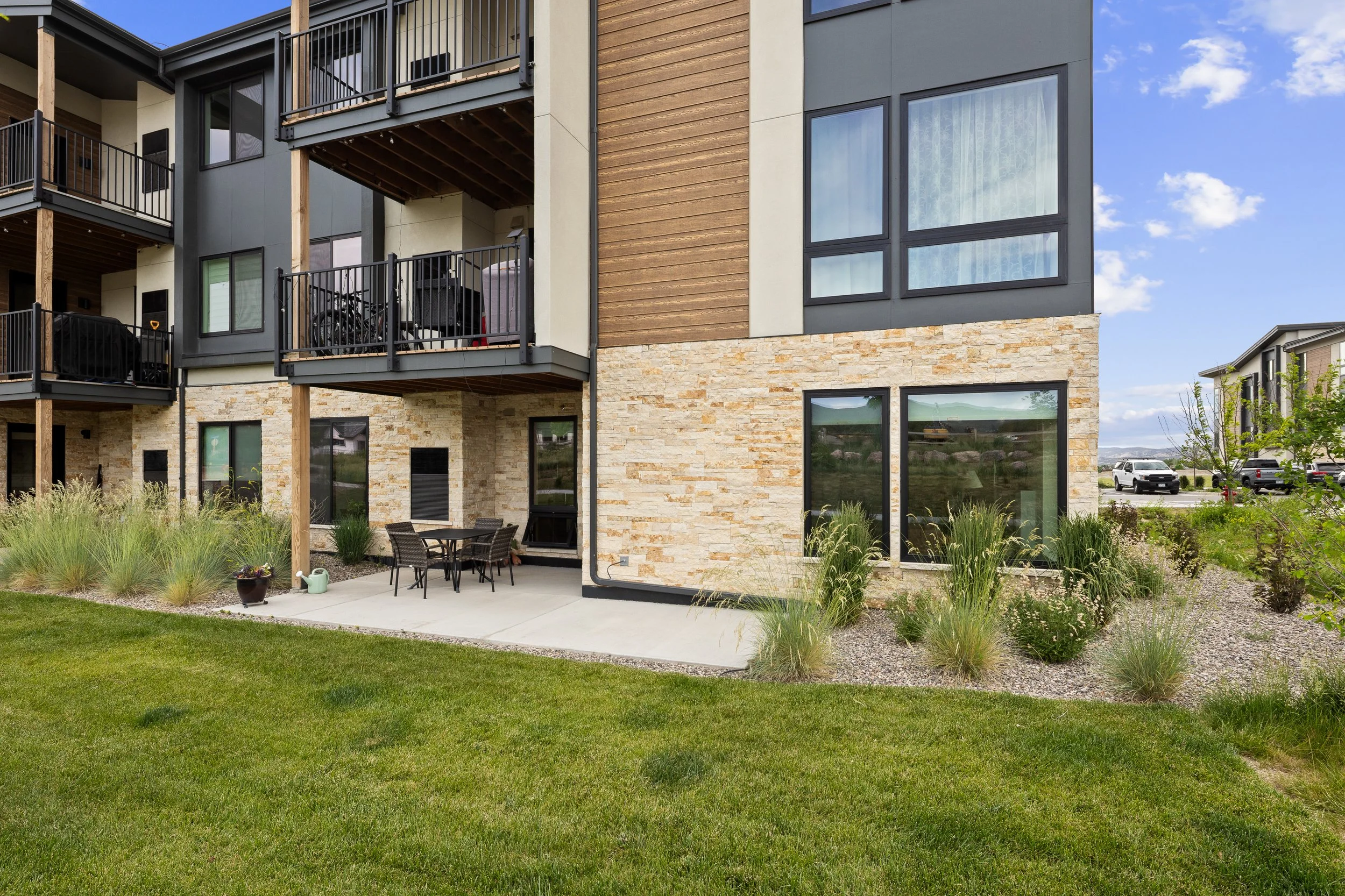 Exterior view of a modern apartment building with balconies, large windows, stone and wood siding, and a landscaped backyard with patio furniture.