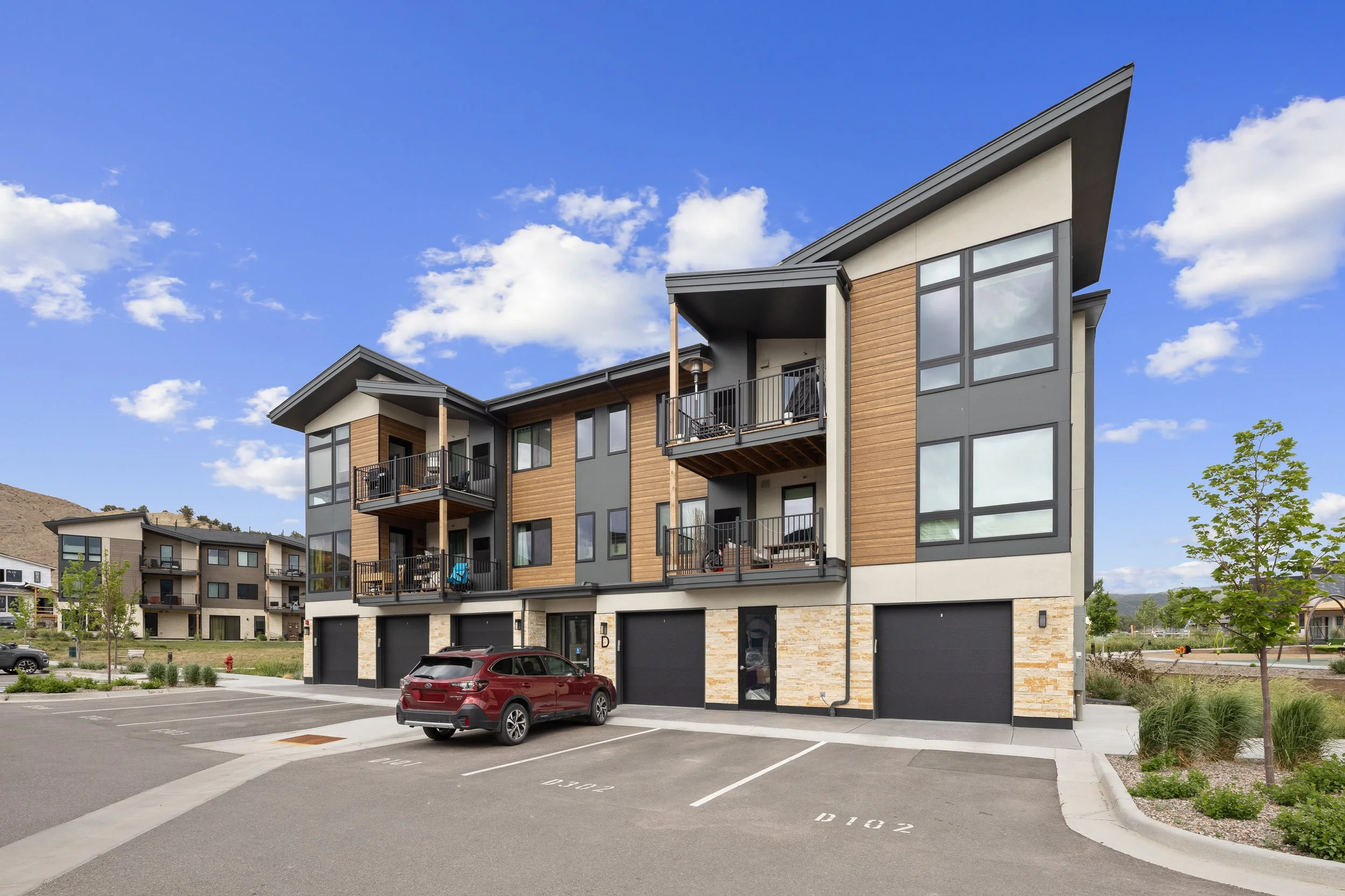 A modern four-story apartment building with wooden and grey siding, large windows, and small balconies, located in a residential area with parking spaces and a red car in front.