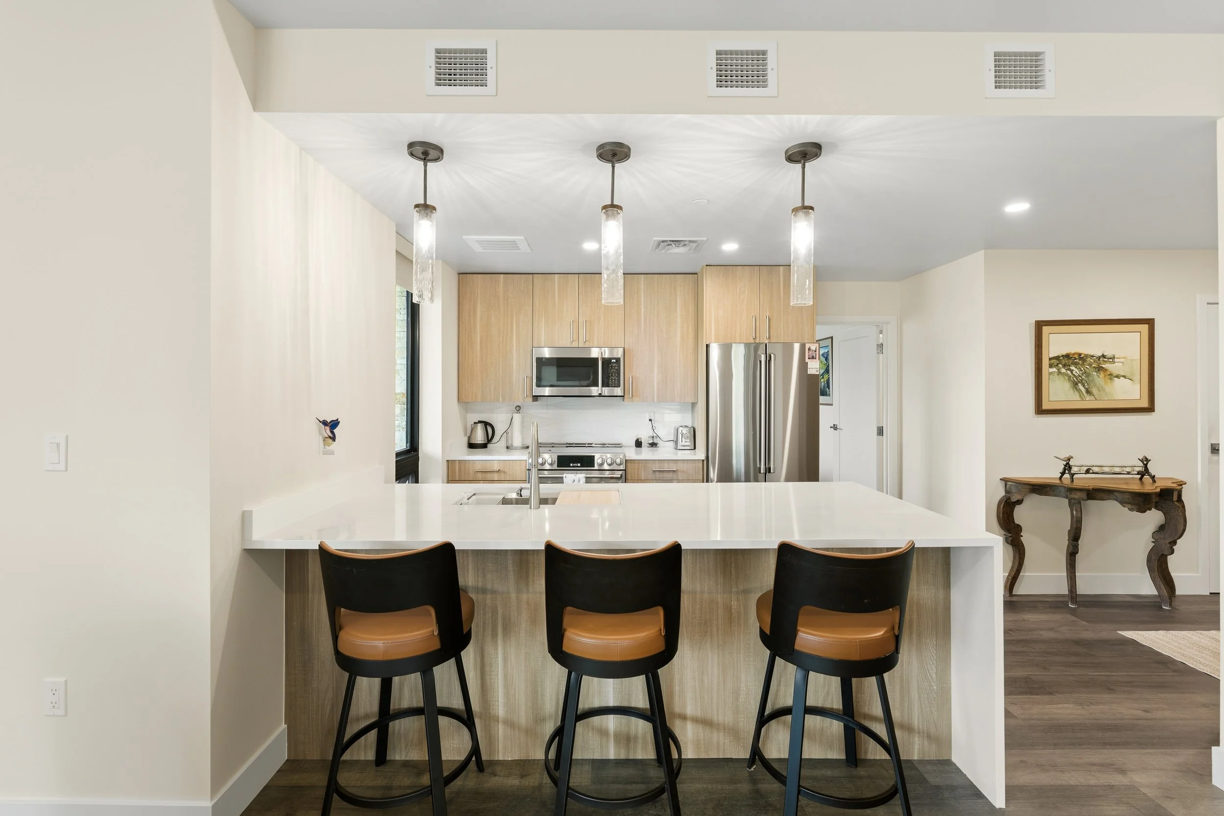 Modern kitchen with beige cabinetry, stainless steel appliances, pendant lighting, black bar stools at a white counter, and artwork on the wall.