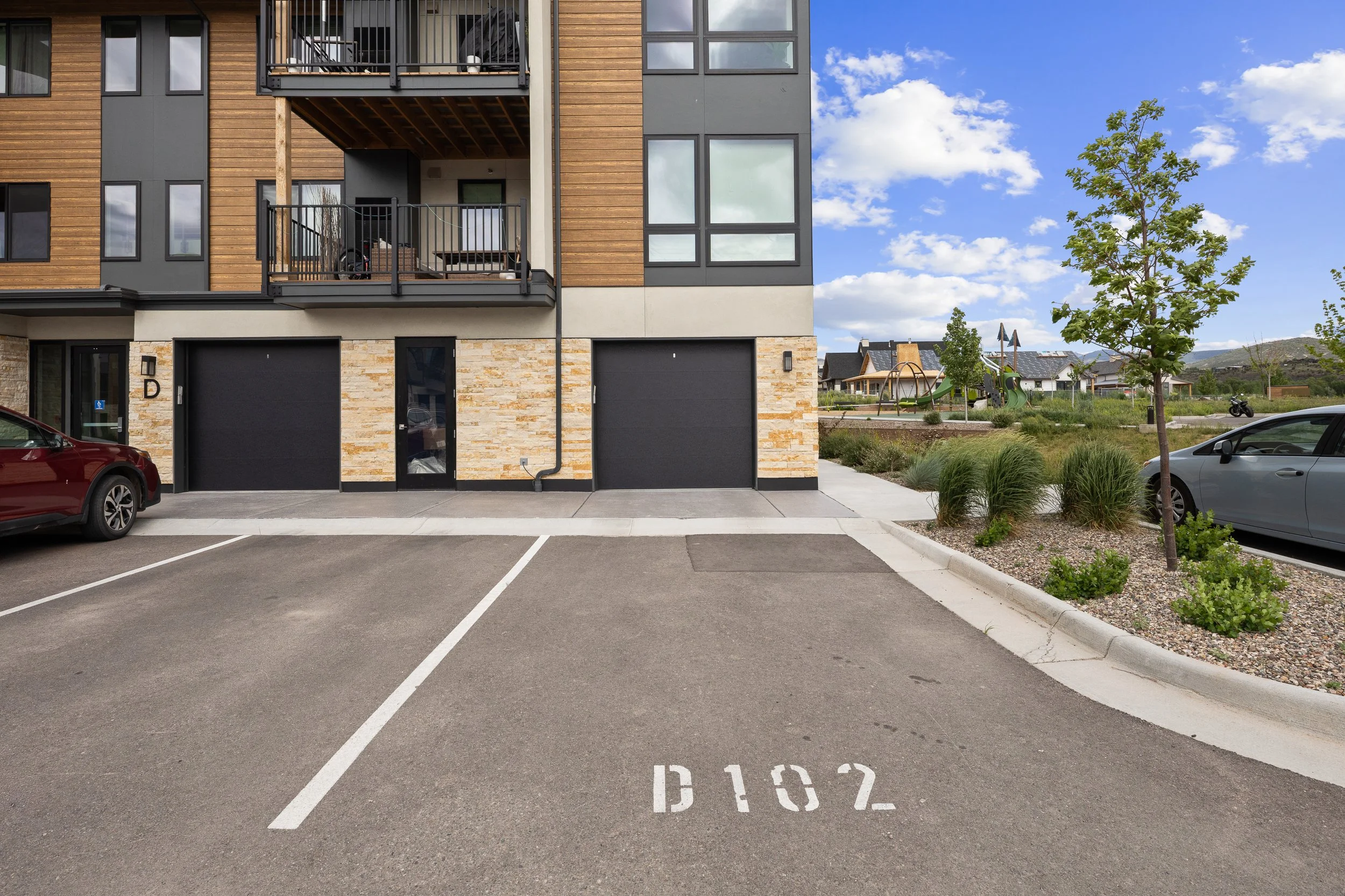 A parking lot in front of a modern apartment building with parking spaces marked B162 and B163. There are two parked cars, one red and one gray, and a small landscaped area with trees and bushes. The building has wood and gray siding with balconies a