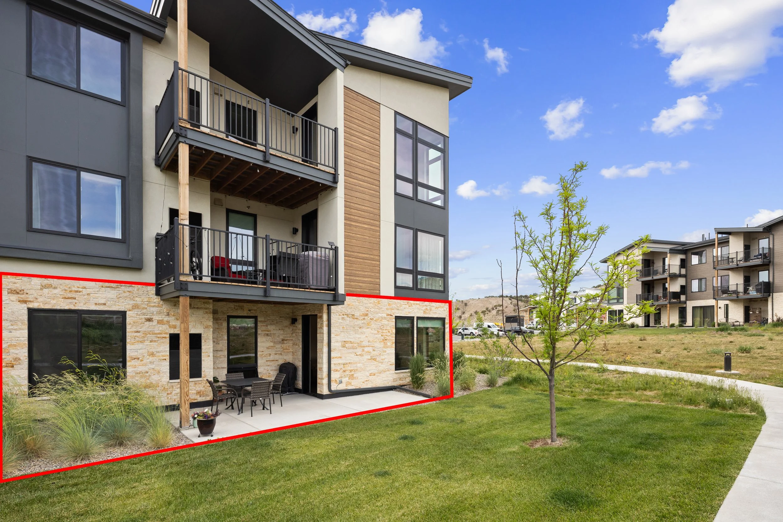 Modern multi-story apartment building with balconies, large windows, and a ground-level patio with outdoor furniture, situated in a landscaped area with a walking path and young trees under a partly cloudy sky.