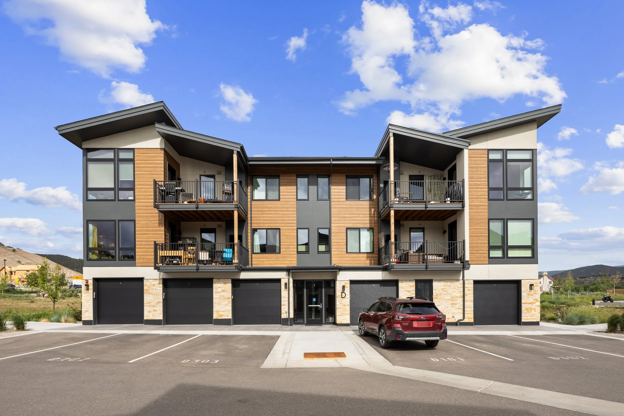 A modern four-story apartment building with a parking lot in front and a mountain landscape in the background.