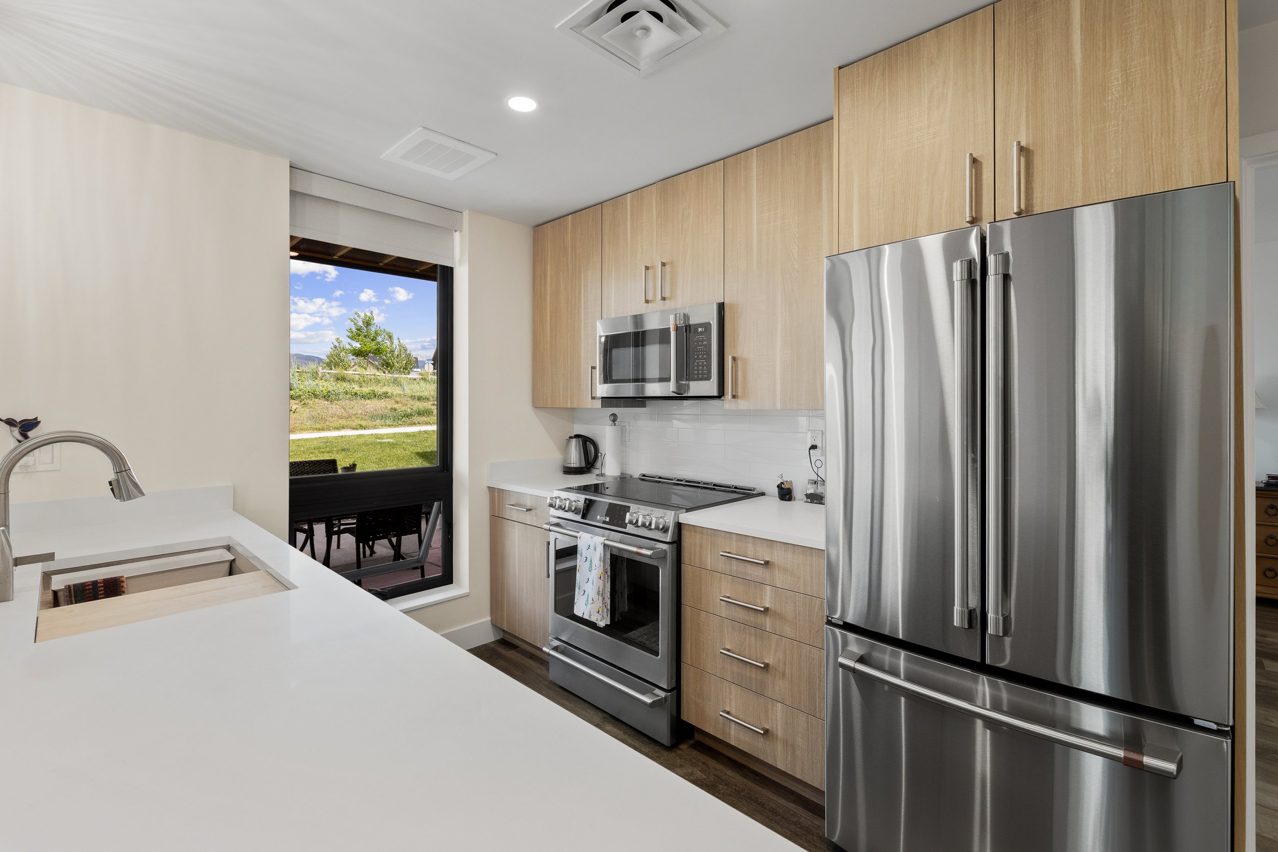 Modern kitchen with wooden cabinets, stainless steel appliances, a microwave, refrigerator, and oven, and a window showing a view of green landscape and blue sky.