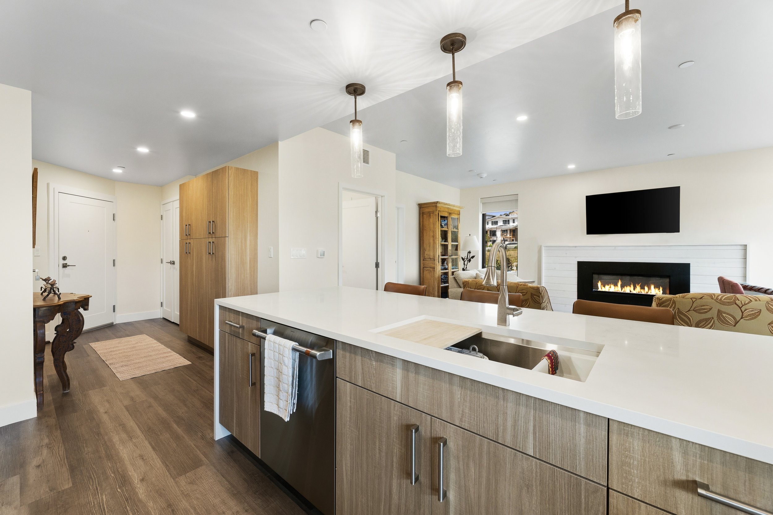 Modern kitchen with white island, wooden cabinets, hardwood floors, and a living room with a fireplace and TV in the background.