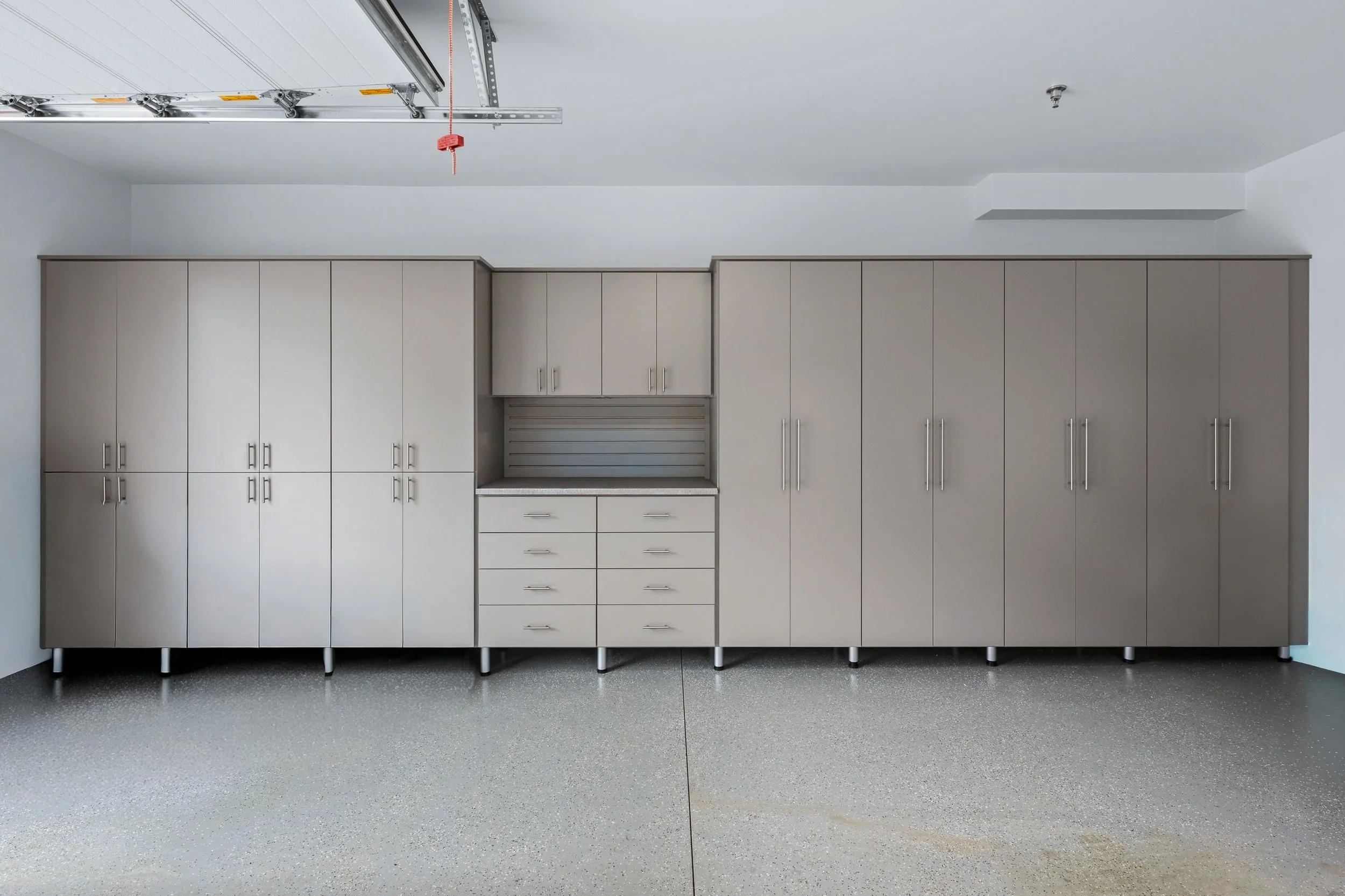 Empty garage with a closed overhead door, white cabinets along the wall, and a gray speckled floor.