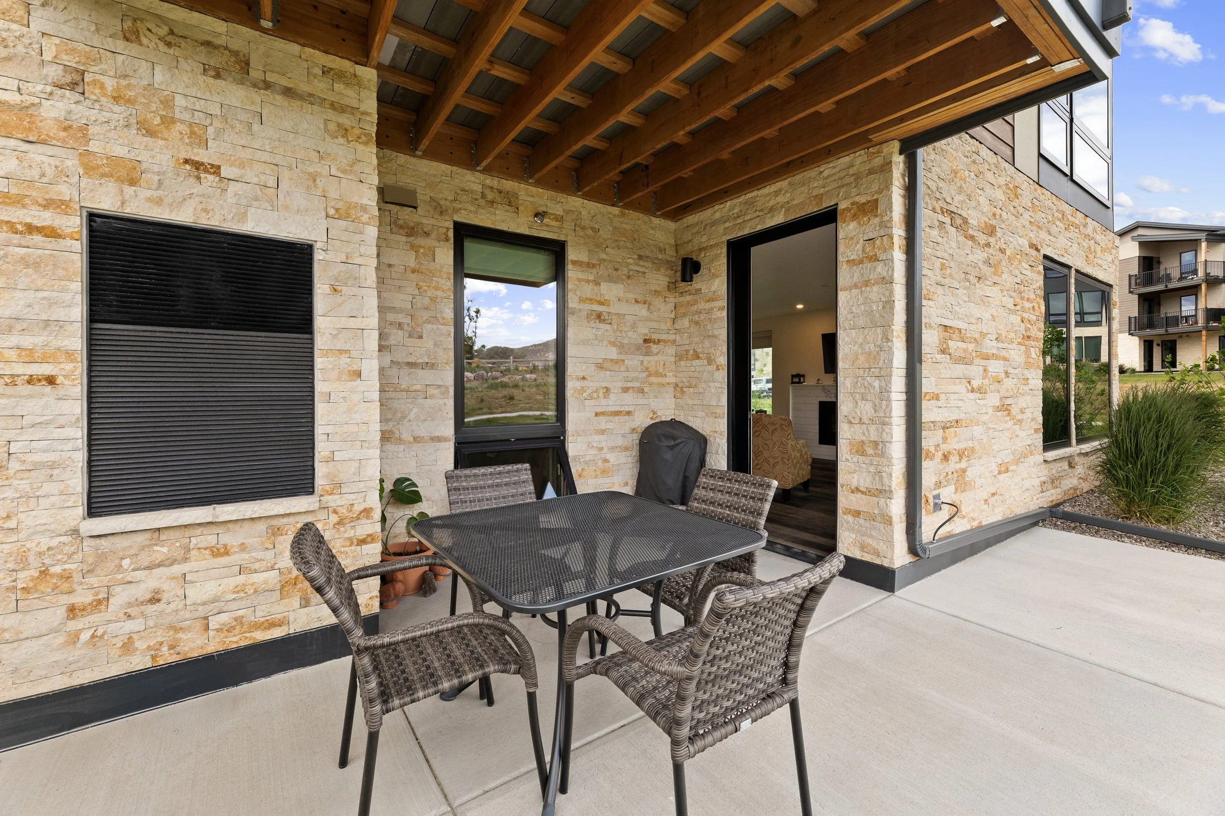 Exterior view of a modern house with stone and black panel siding, a covered porch with patio furniture, and scenic landscape with apartment buildings in the background.