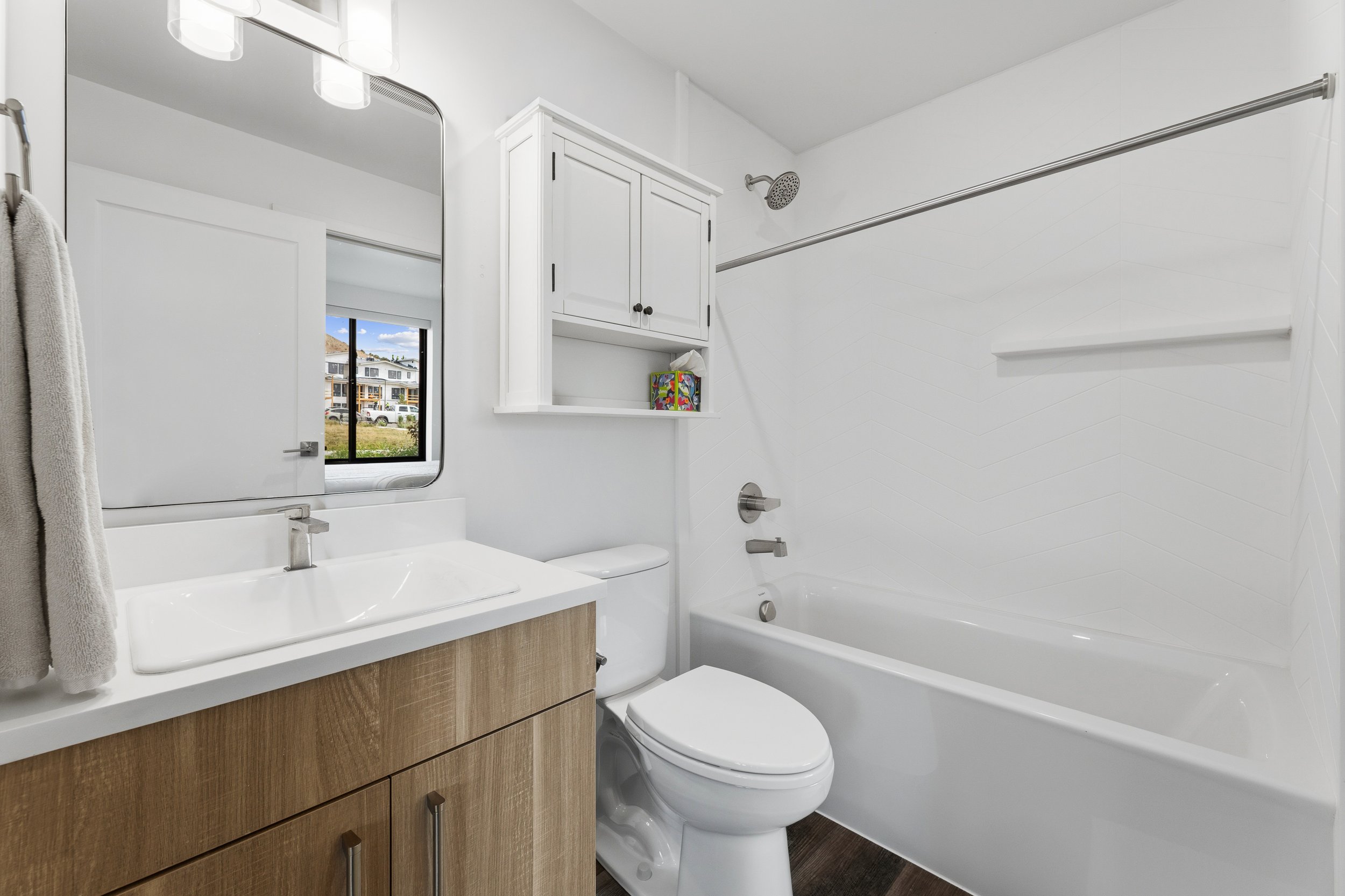 A modern white bathroom with a mirror above a sink, a cabinet, a toilet, and a bathtub with a shower, a window showing an outside view of buildings and a grassy area.