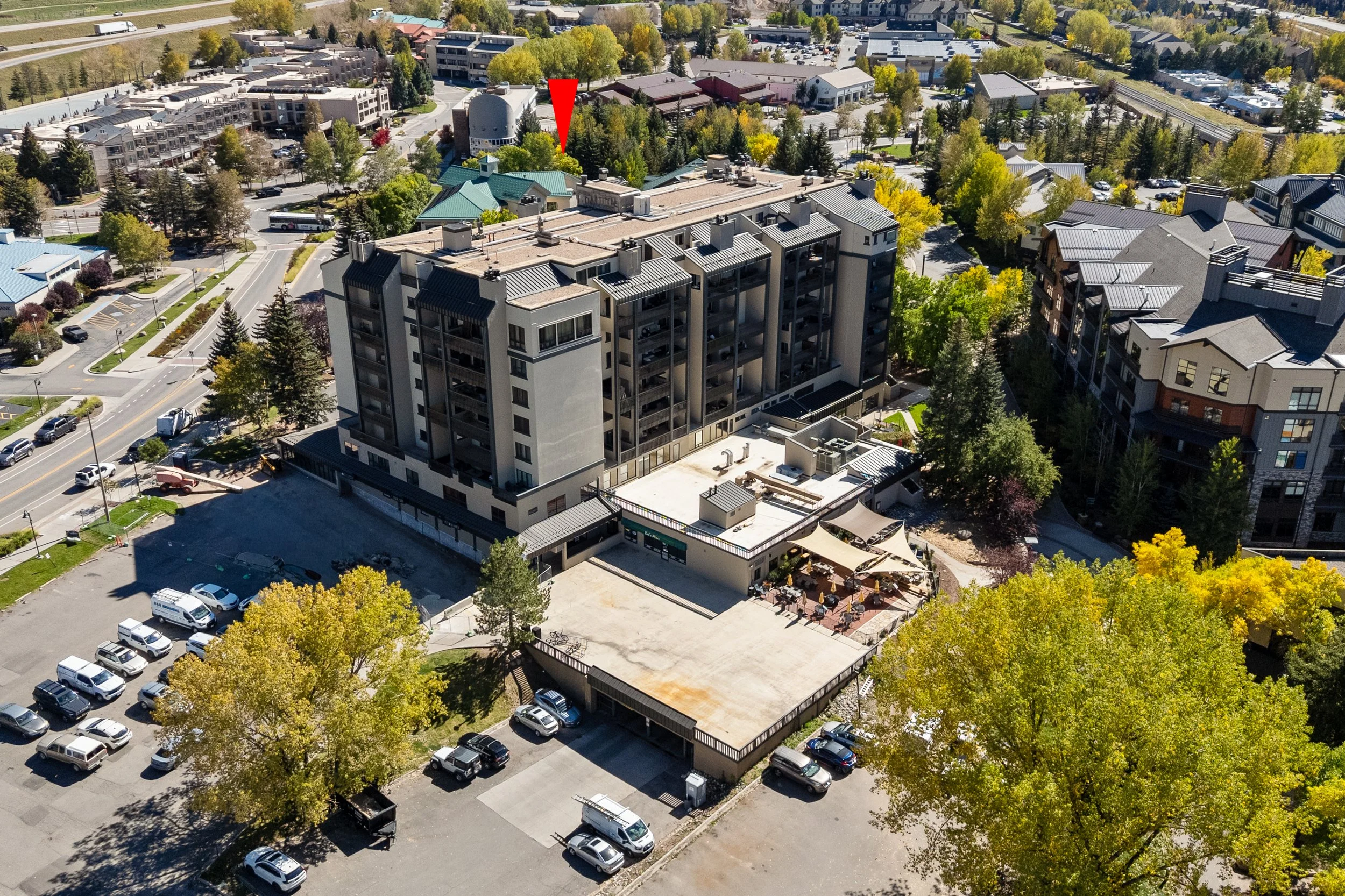 An aerial view of a cityscape featuring a large multi-story building with outdoor seating on a terrace, surrounded by trees with autumn foliage, parking lots with many cars, and smaller buildings with varied rooftops.