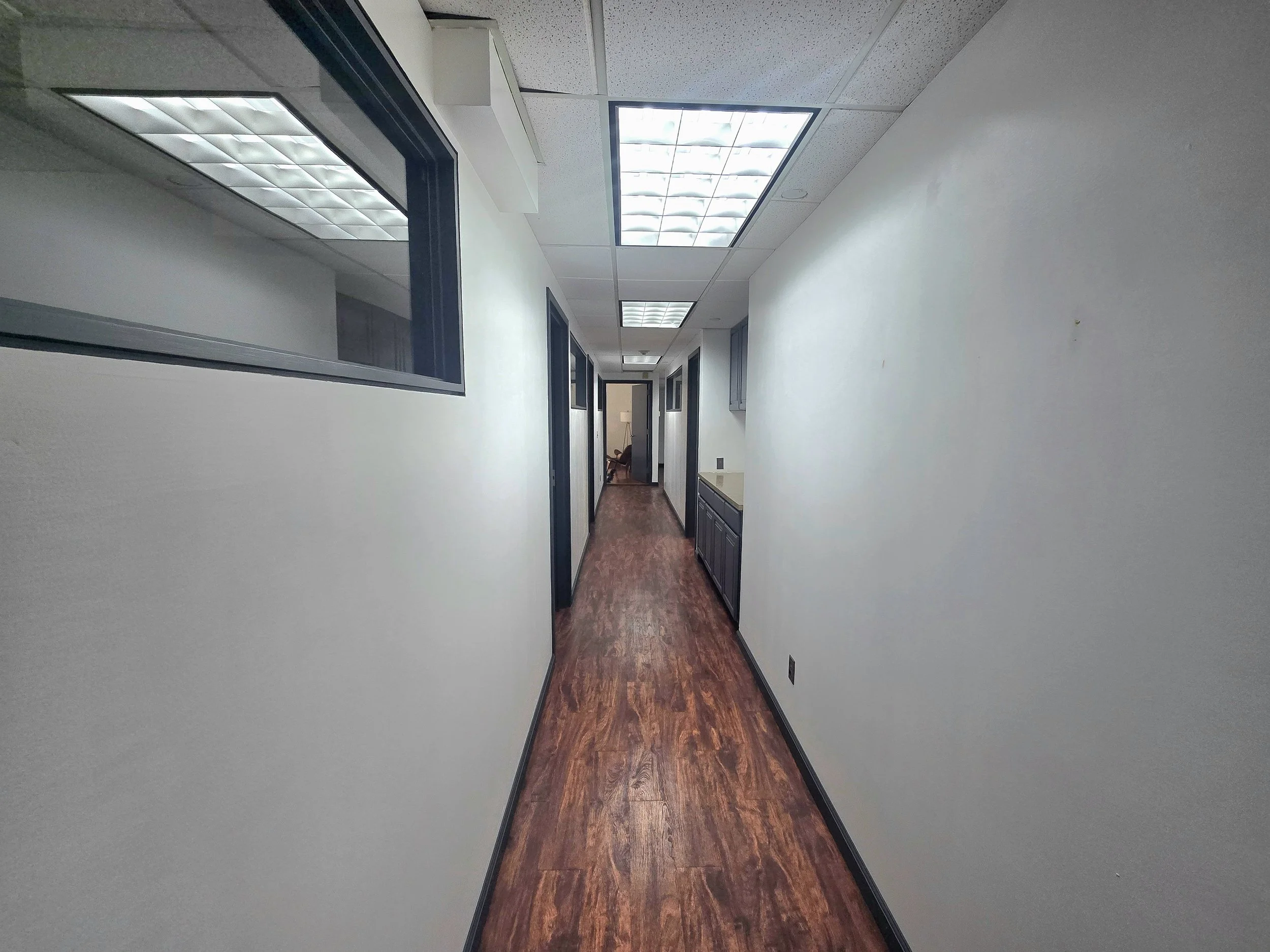 A narrow hallway with white walls, wooden flooring, window and door frames, and ceiling lights.