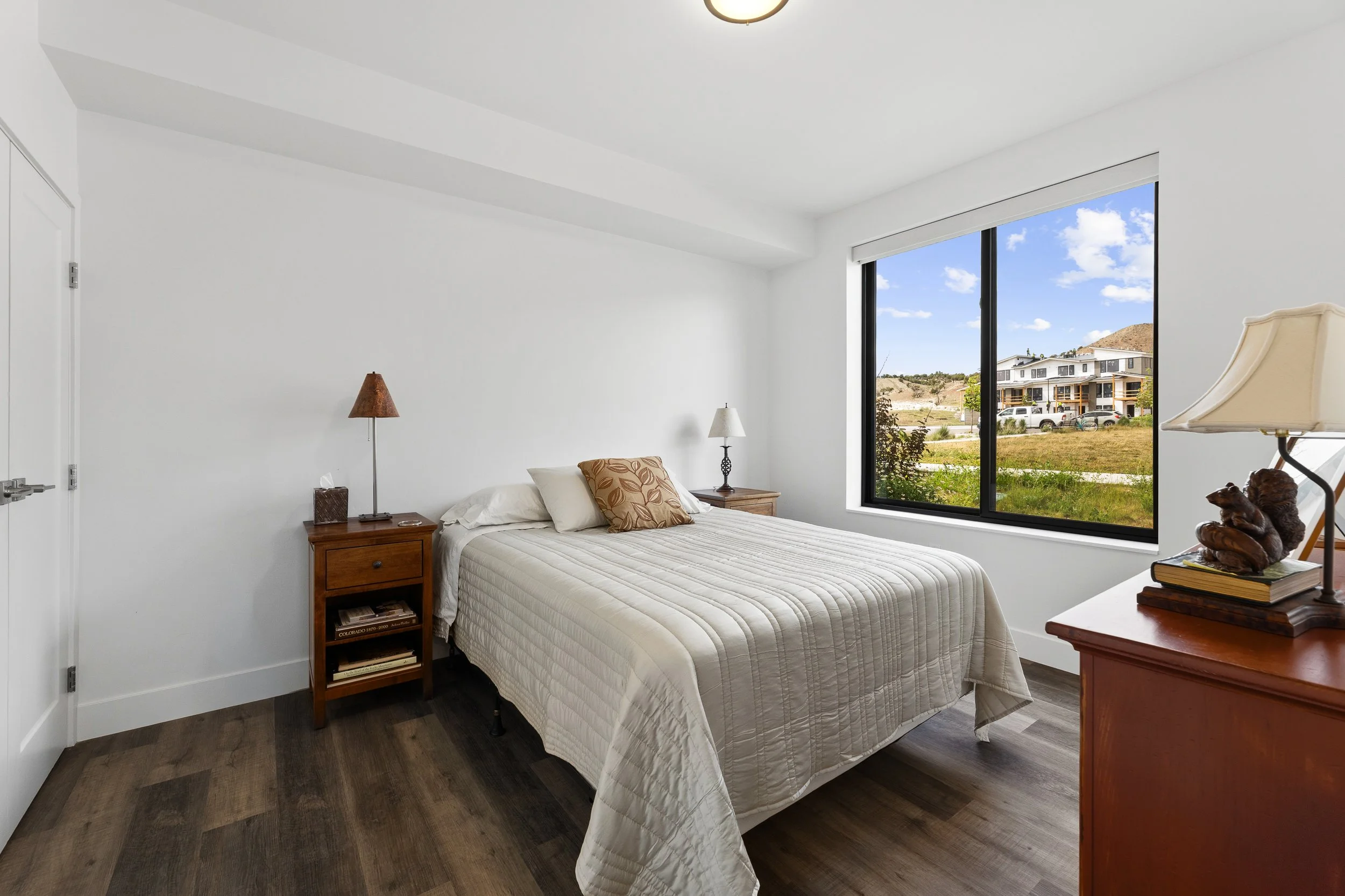Bedroom with a bed, wooden nightstand, lamp, window showing outdoor houses and a partly cloudy sky.