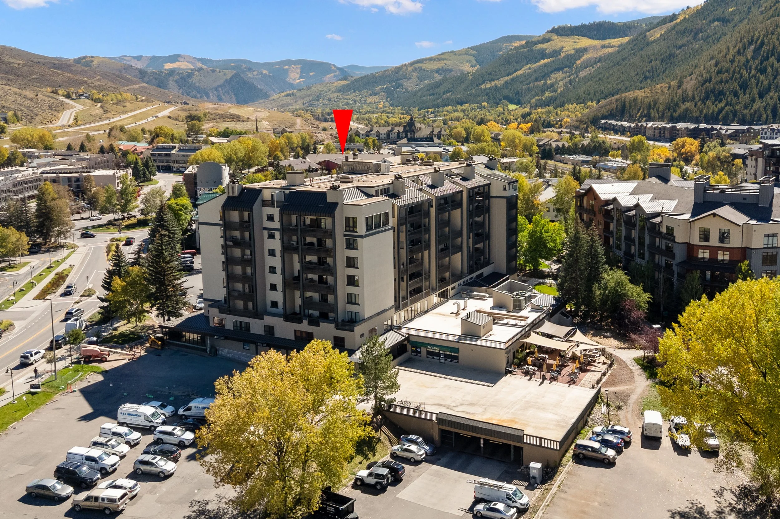 A daytime aerial view of a mountain town with multi-story residential and commercial buildings, parking lots filled with cars, trees with fall foliage, and a backdrop of green mountains and blue sky.