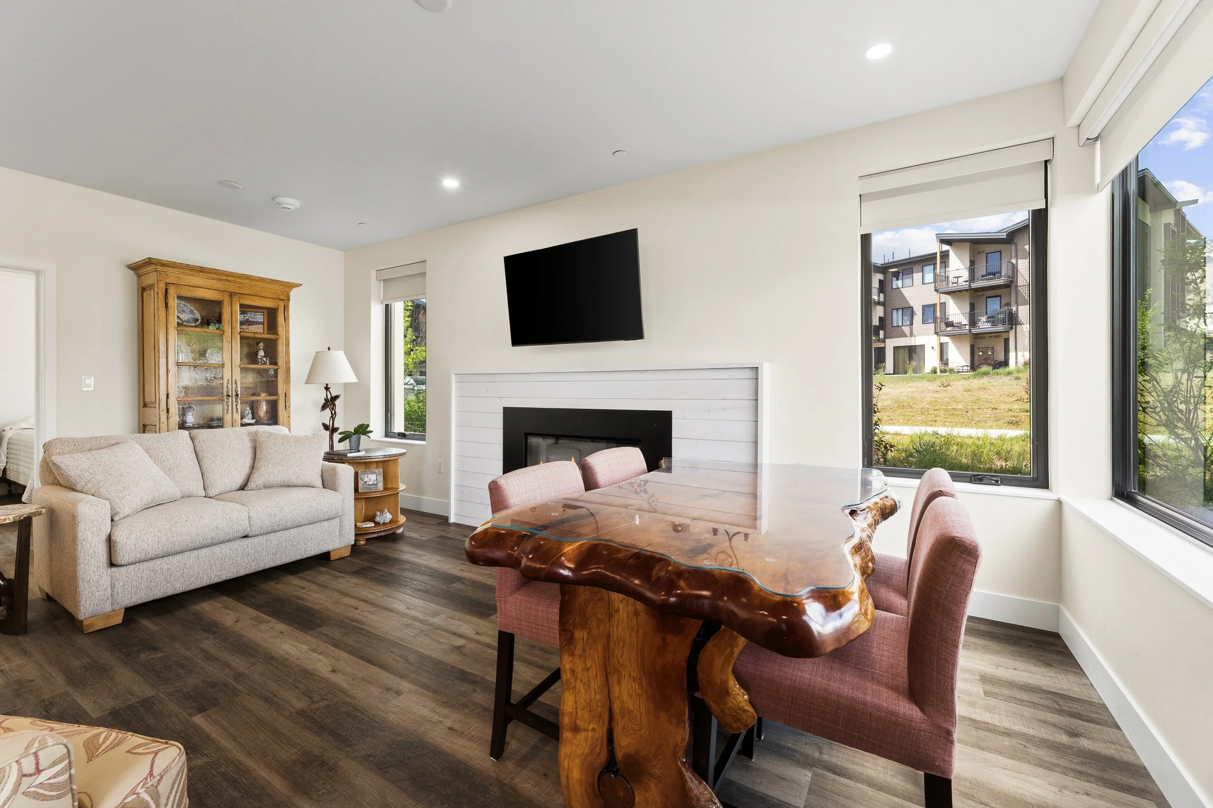 A living room with a beige couch, a wooden dining table with pink upholstered chairs, a white fireplace, a wooden cabinet, a side table with a lamp, and large windows showing an apartment building outside.