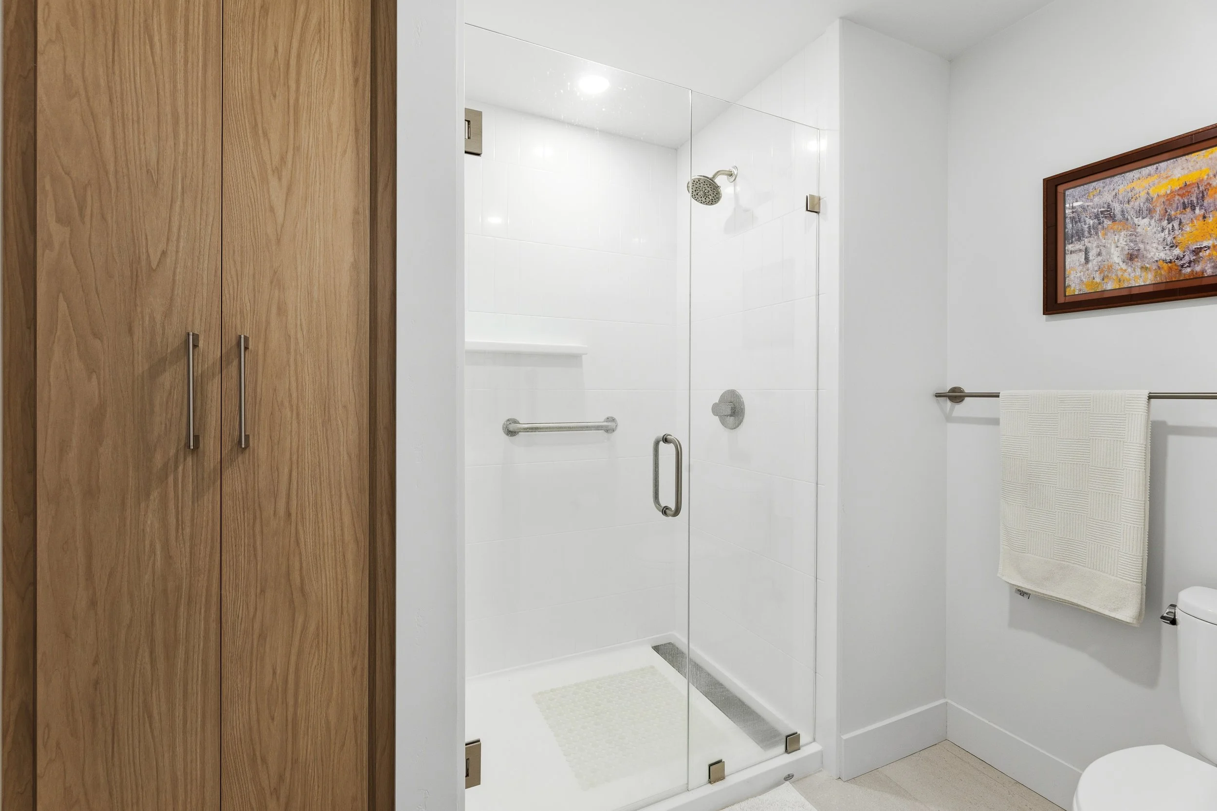 Clean bathroom with white walls, glass shower enclosure, wooden cabinet on the left, a towel rack with a white towel, a framed painting on the wall, and a toilet partially visible.