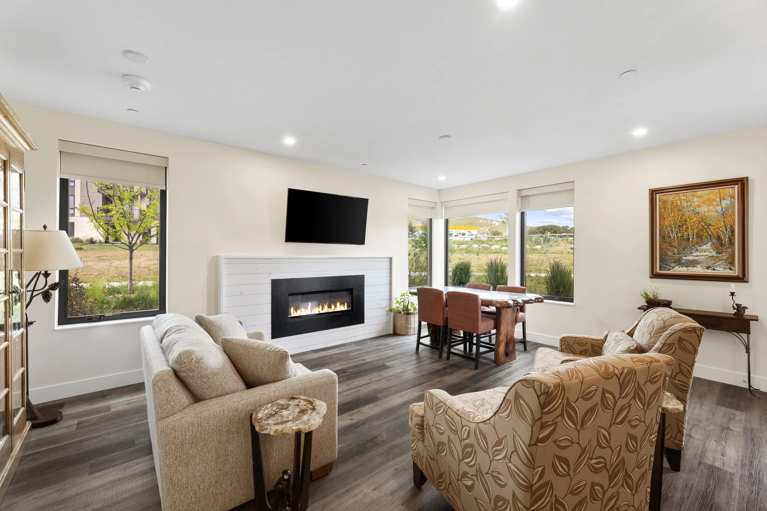 Living room with large windows, a fireplace, a wall-mounted TV, a dining table with chairs, sofas, a painting, and decorative plants.